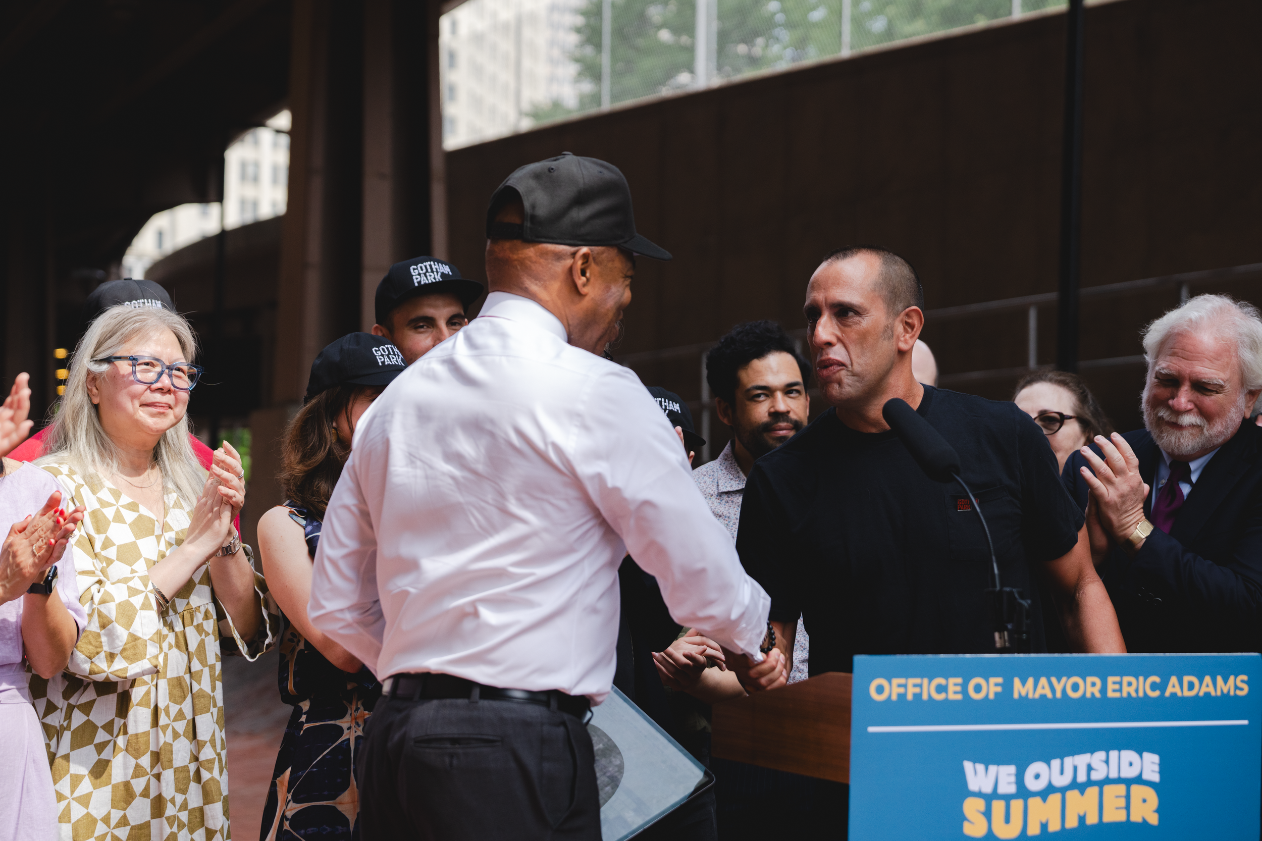 Group of diverse people gathered outdoors, some wearing 'Gotham Park' hats, with two men shaking hands at a podium labeled 'Office of Mayor Eric Adams,' during a summer event. The Banks. Gotham Park Opening. Eric Adams