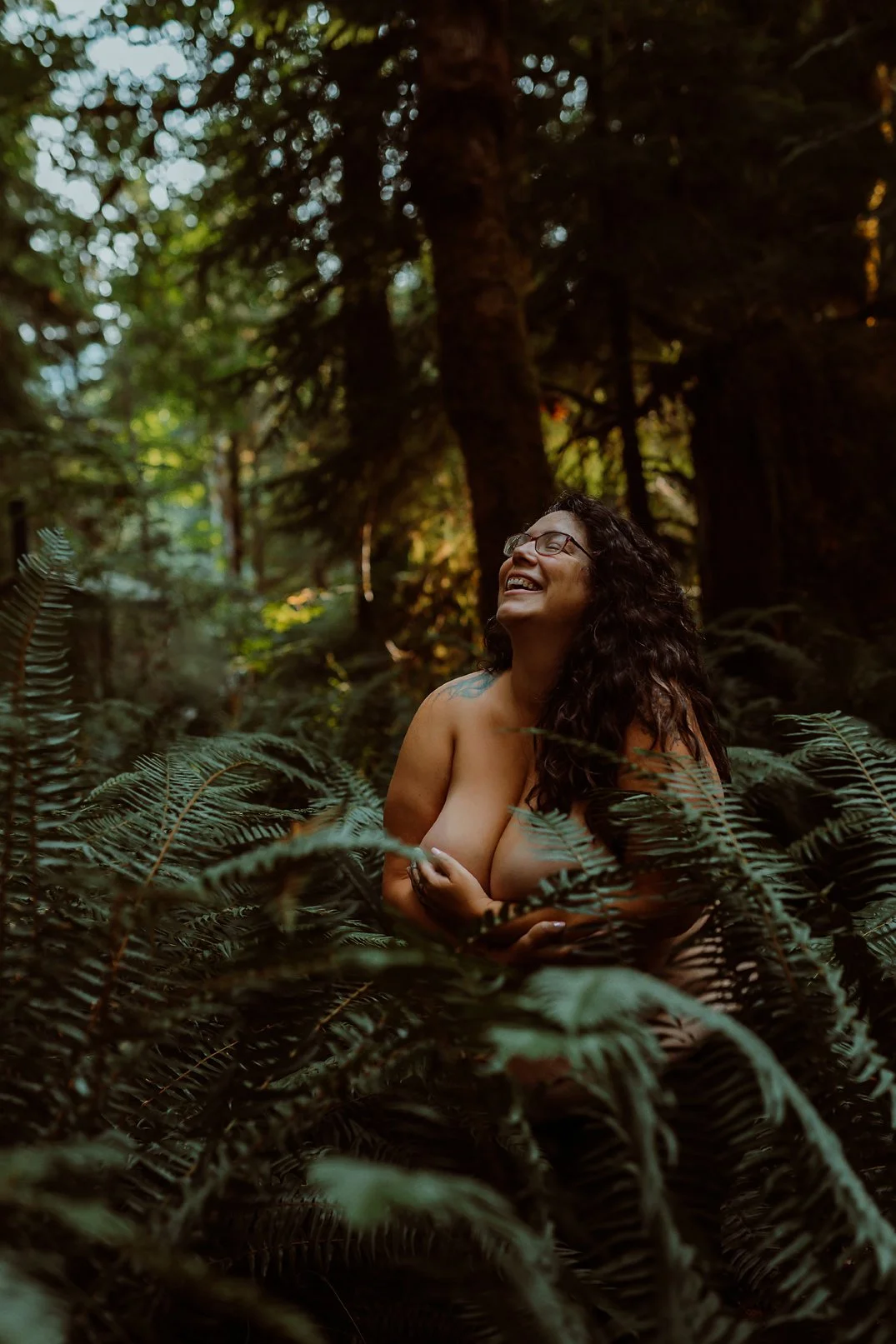 A joyful woman with curly hair and glasses is smiling and sitting amidst lush green ferns in a forest, with tall trees and dappled sunlight in the background.