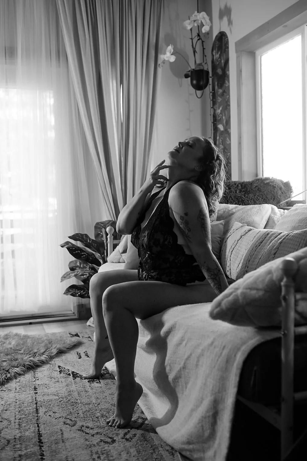 A woman with tattoos sitting on a sofa in a living room, dressed in lace lingerie, posing with her head tilted back and hand on her neck, in black and white.