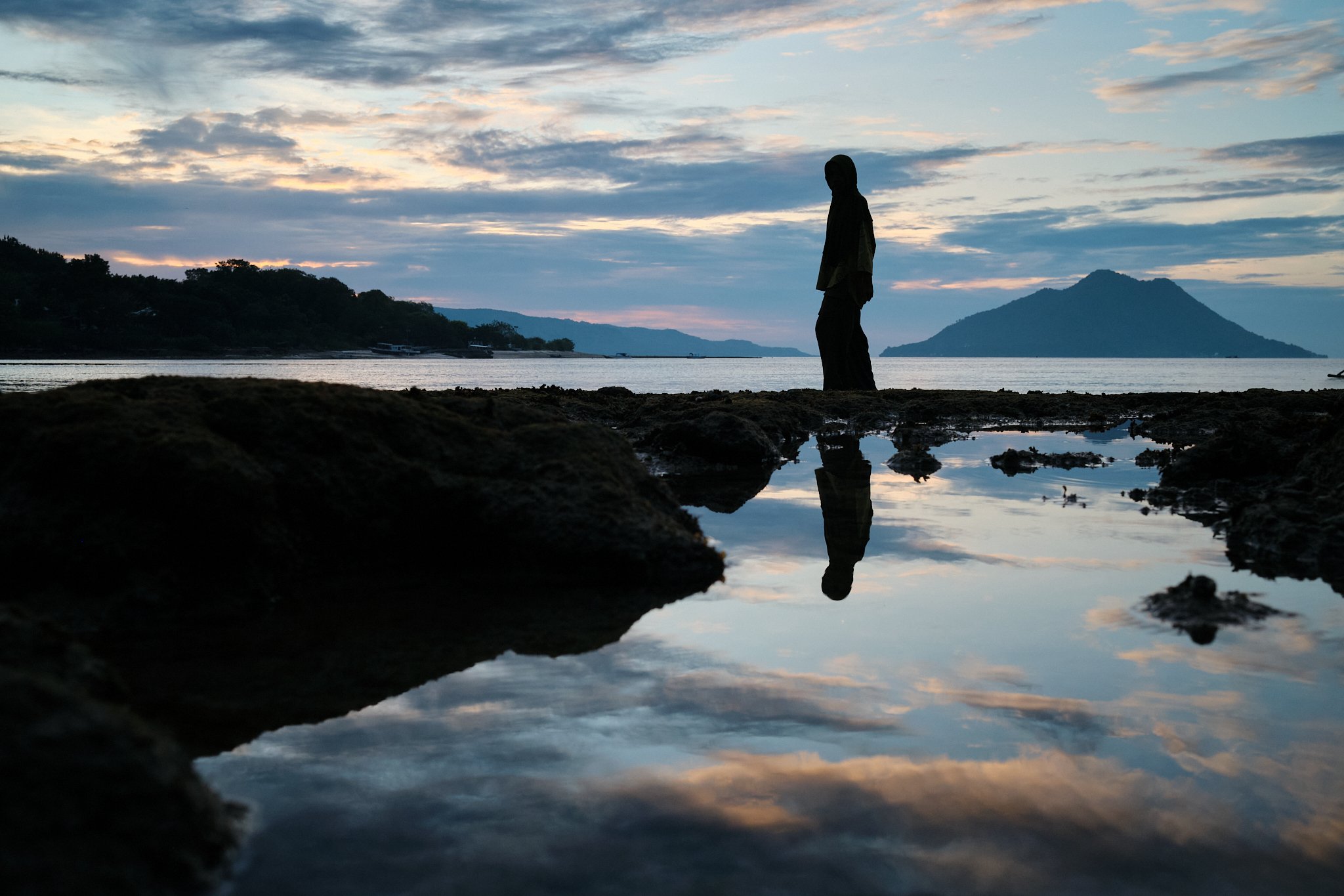 A woman in silhouette walks along a rocky shoreline at dusk in Alor, Indonesia, her reflection mirrored in a tide pool below. Photo by travel photographer Lee Starnes.