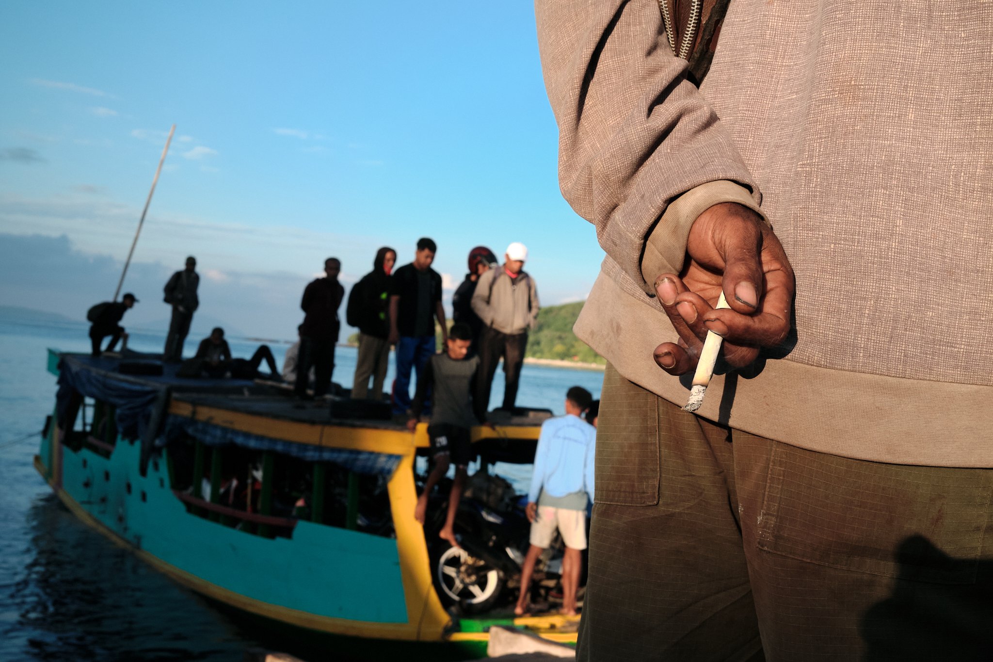 Close-up of a man holding a cigarette, while a group of people stands on a turquoise wooden boat in the background. Captured in Alor, Indonesia by travel photographer Lee Starnes.