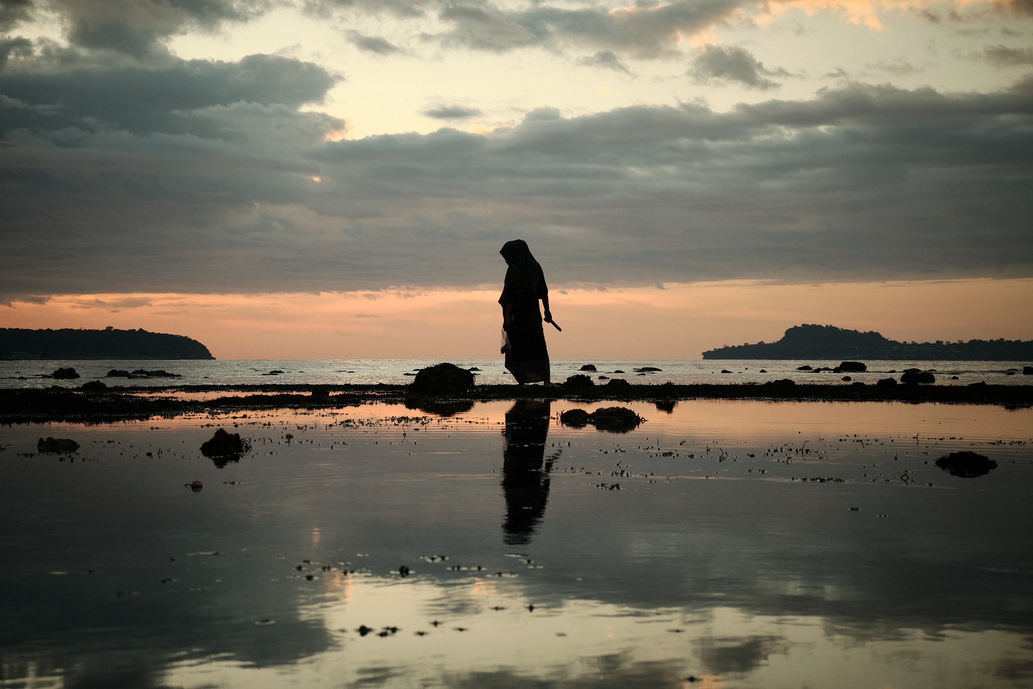 A solitary woman in traditional clothing walks through a shallow tidal flat at sunset, casting a striking reflection in the still water. Taken in Alor, Indonesia by travel photographer Lee Starnes.