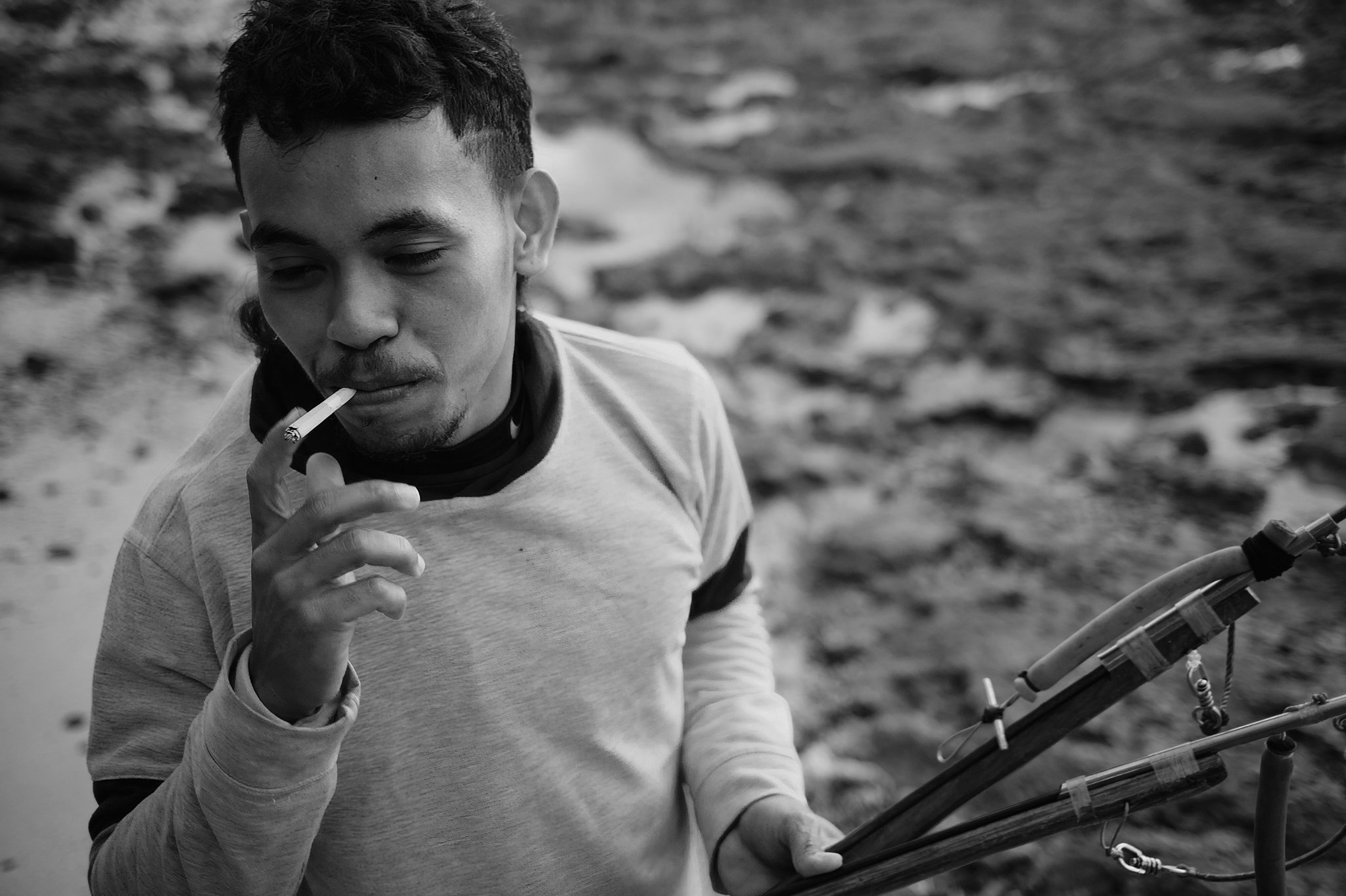 A black-and-white image of a young man lighting a cigarette while holding fishing gear, set against the rugged shoreline of Alor, Indonesia. Photo by Lee Starnes.