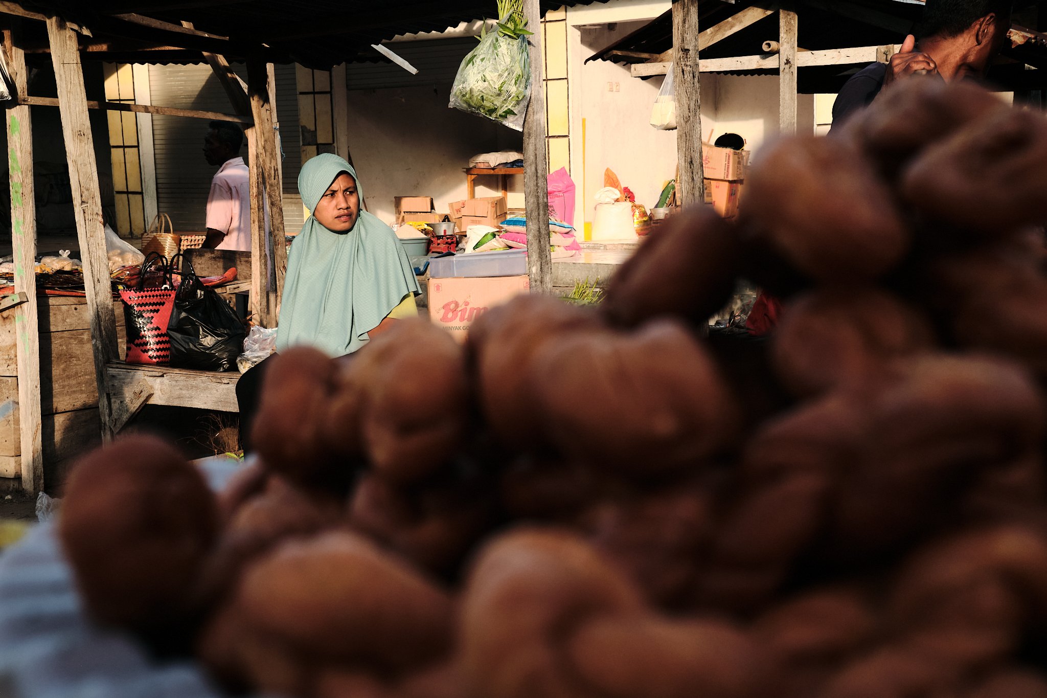 A woman in a mint green headscarf sits behind a market stall, framed by soft morning light and local produce in a rustic setting. Taken in Alor, Indonesia by travel photographer Lee Starnes.