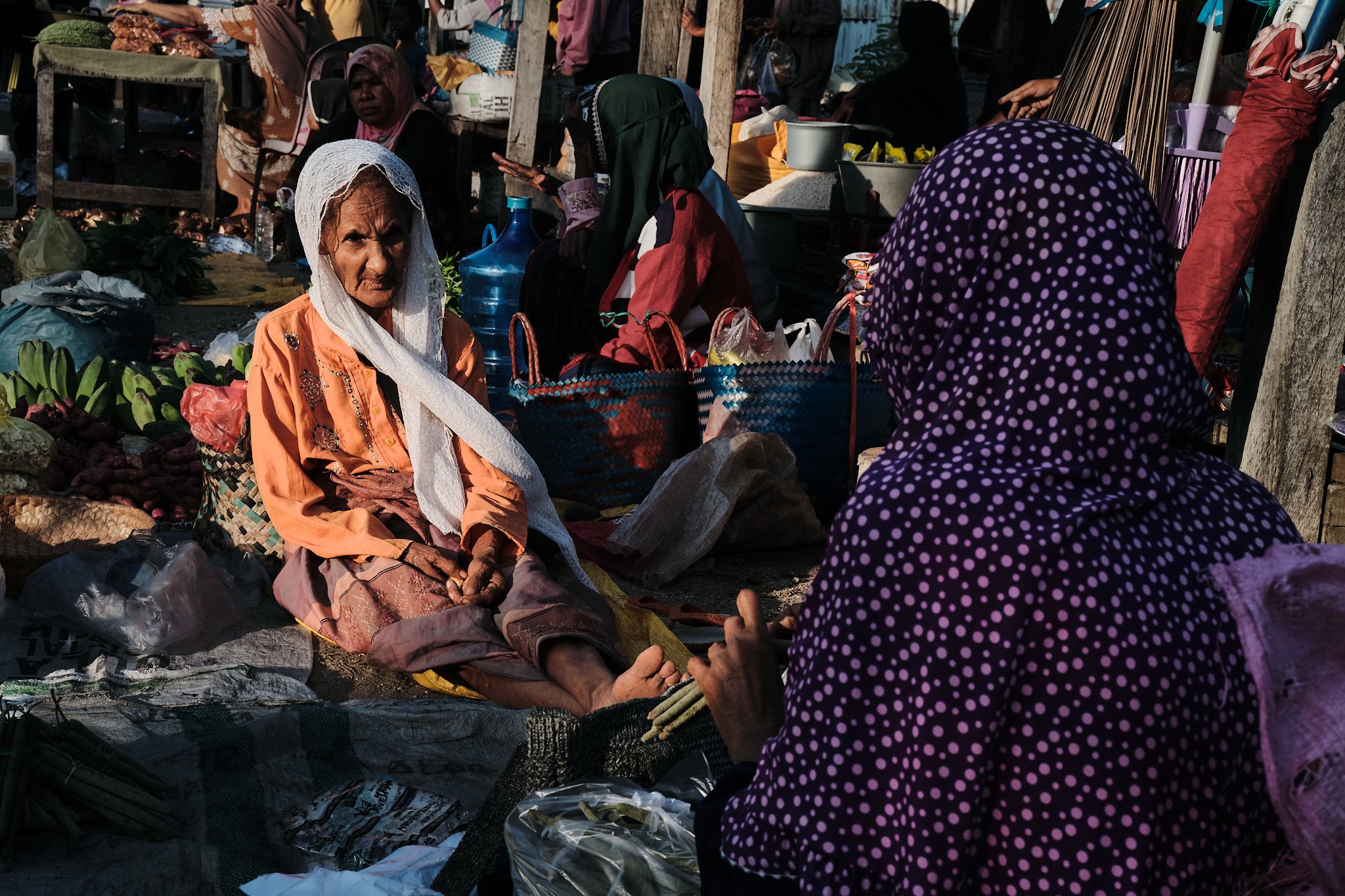An elderly woman in an orange blouse and white scarf sits barefoot on a cloth in a crowded outdoor market, engaged in conversation. Photographed in Alor, Indonesia by Lee Starnes.