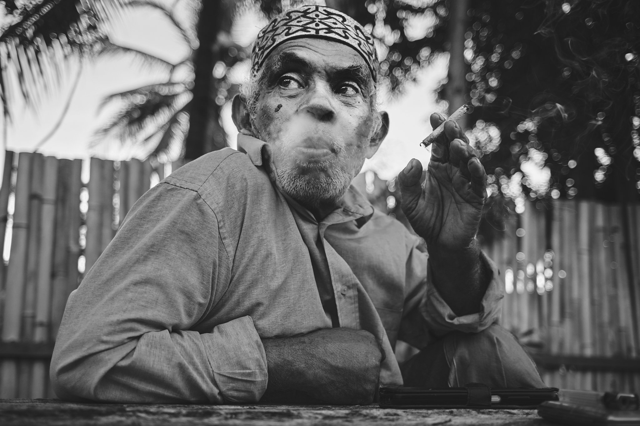 Black-and-white portrait of an older man with a patterned cap exhaling smoke from a cigarette, seated in front of a bamboo fence under palm trees. Taken in Alor, Indonesia by travel photographer Lee Starnes.