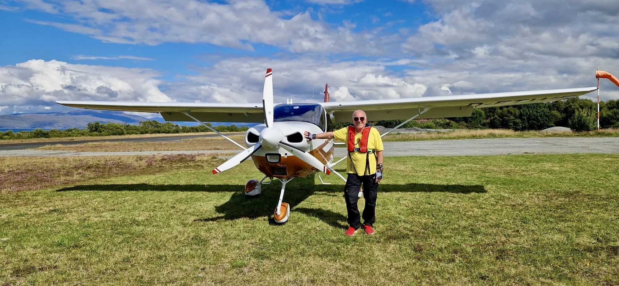 Ein Mann steht neben einem kleinen Flugzeug auf einem Flugfeld, trägt eine gelbe T-Shirt, eine rote Schwimmweste und Sonnenbrille, bei sonnigem Himmel mit Wolken im Hintergrund.