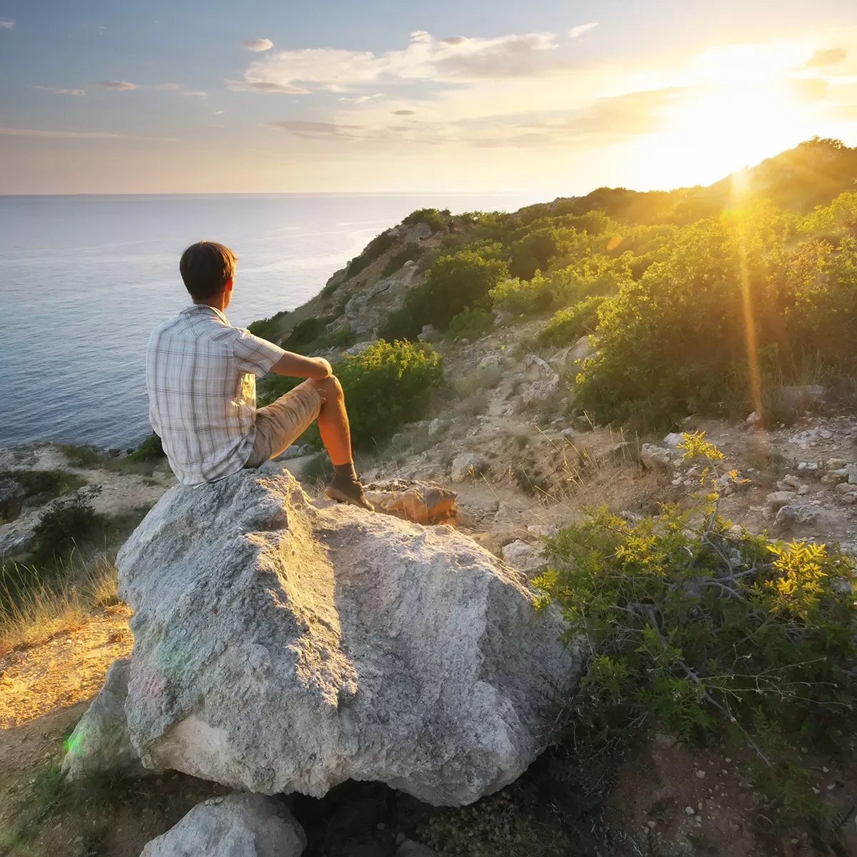 Ein Junge sitzt auf einem großen Felsen und betrachtet die untergehende Sonne über dem Meer, umgeben von einer hügeligen Landschaft mit Büschen und Bäumen.