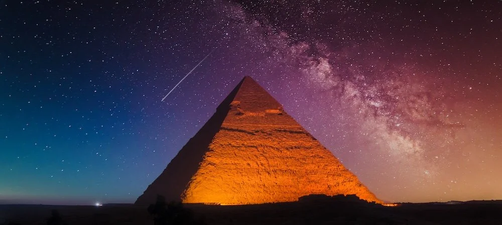Night sky with stars and the Milky Way galaxy over a pyramid illuminated by orange lights.