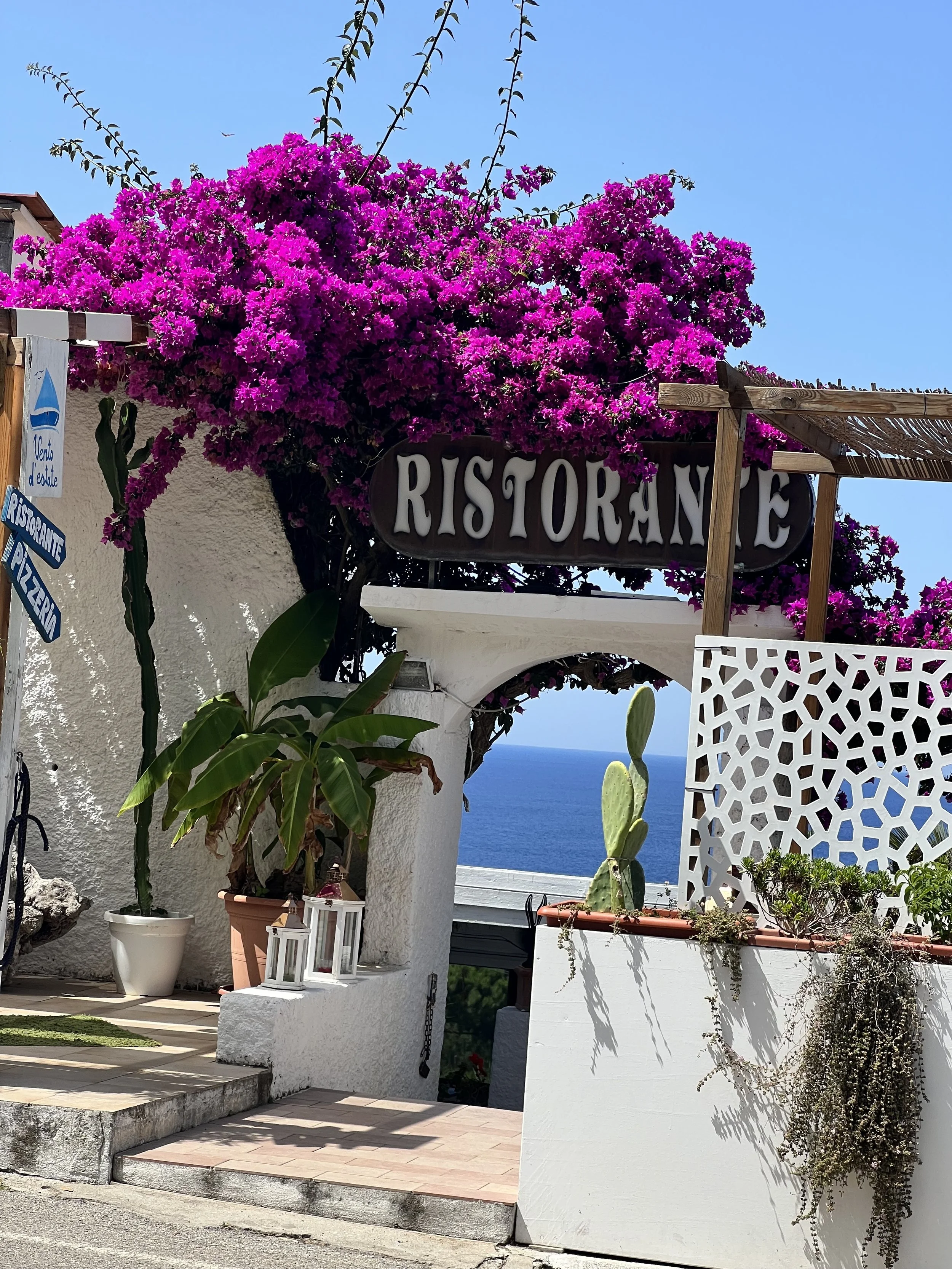 Blick auf einen mediterranen Küstenrestaurant-Eingang mit Bougainvillea-Blumen und Meer im Hintergrund.