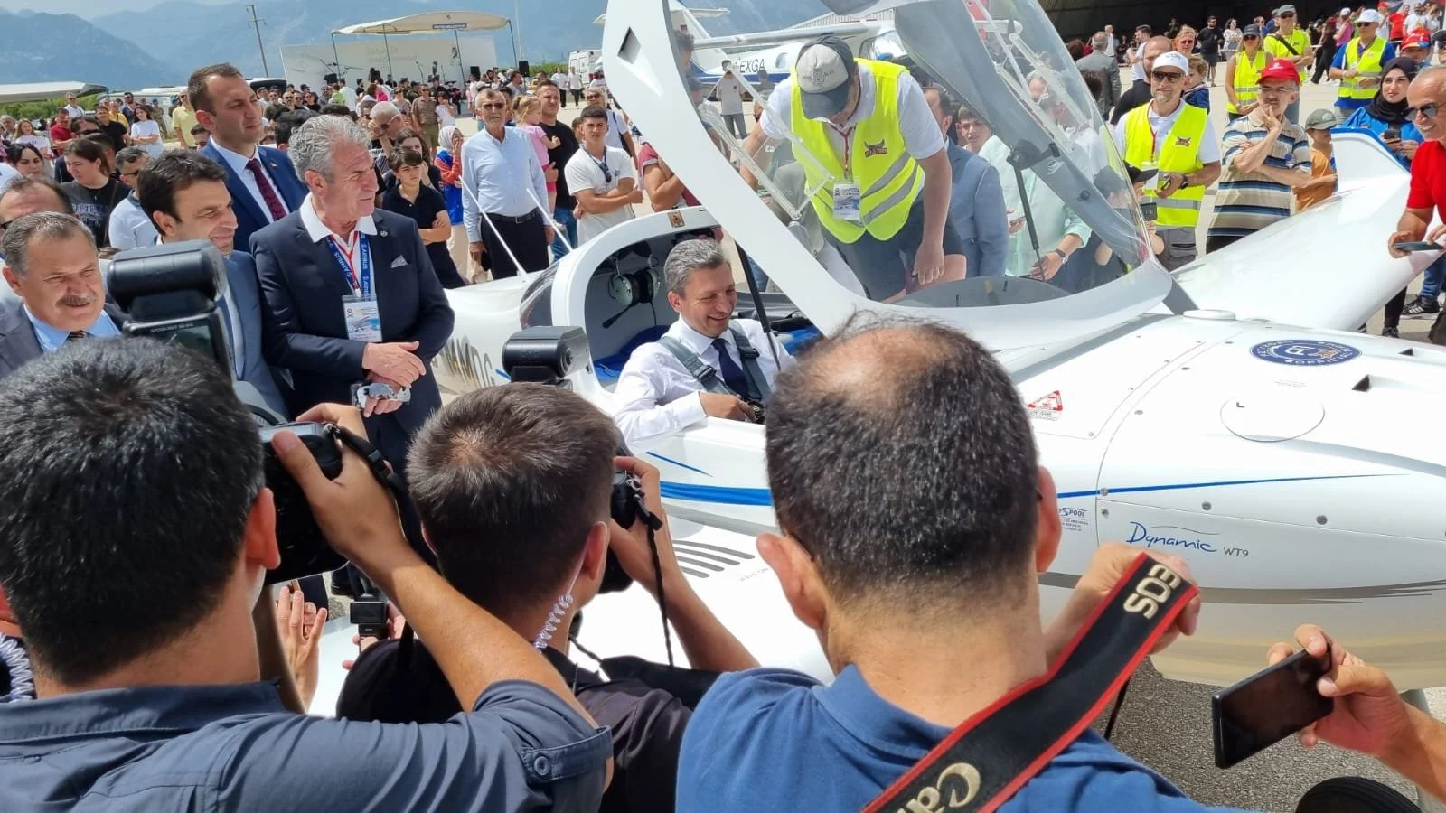 Ein Mann sitzt in einem weißen, futuristischen Flugzeug, umgeben von einer Menschenmenge, die das Flugzeug begutachtet. Einige Personen nehmen Fotos auf, während andere das Geschehen beobachten.