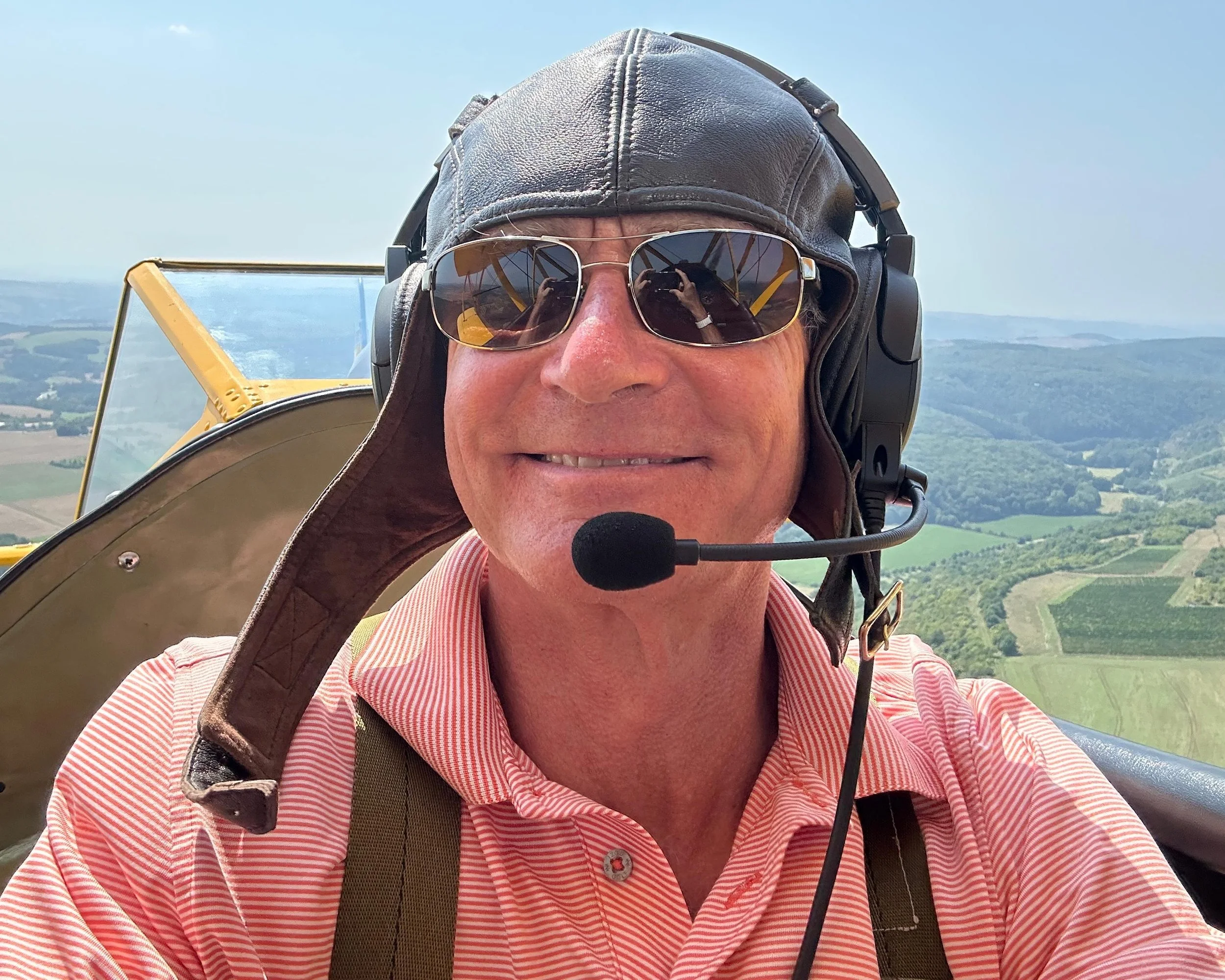 Ein Mann fliegt in einem gelben Doppeldecker-Flugzeug, trägt eine Lederkappe, Sonnenbrille und ein Headset, mit einer Landschaft im Hintergrund.