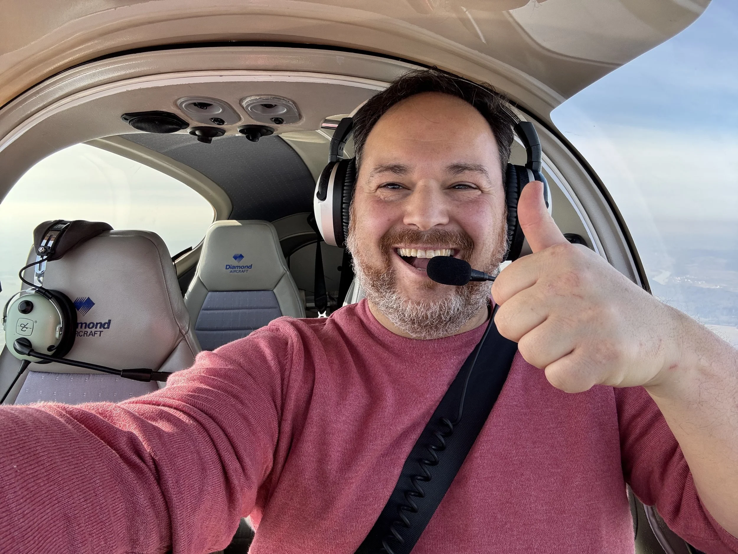 Ein glücklicher Mann im Cockpit eines Kleinflugzeugs, trägt ein Headset mit Mikrofon, macht einen Daumen hoch und lächelt in die Kamera.