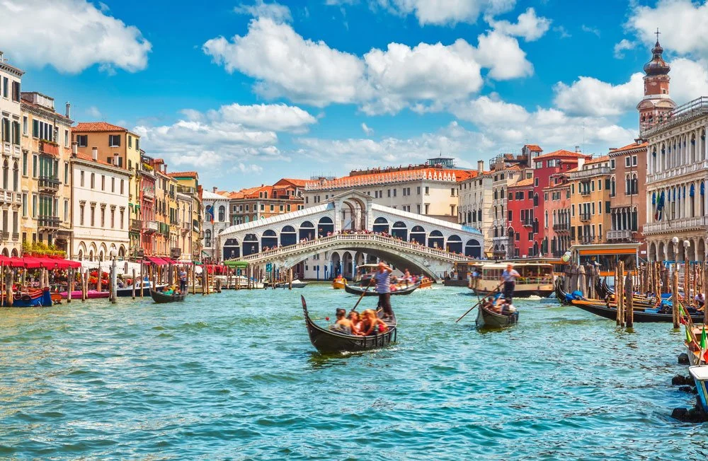 A view of the Grand Canal in Venice, Italy, with gondolas, colorful historic buildings, and the Rialto Bridge under a partly cloudy sky.