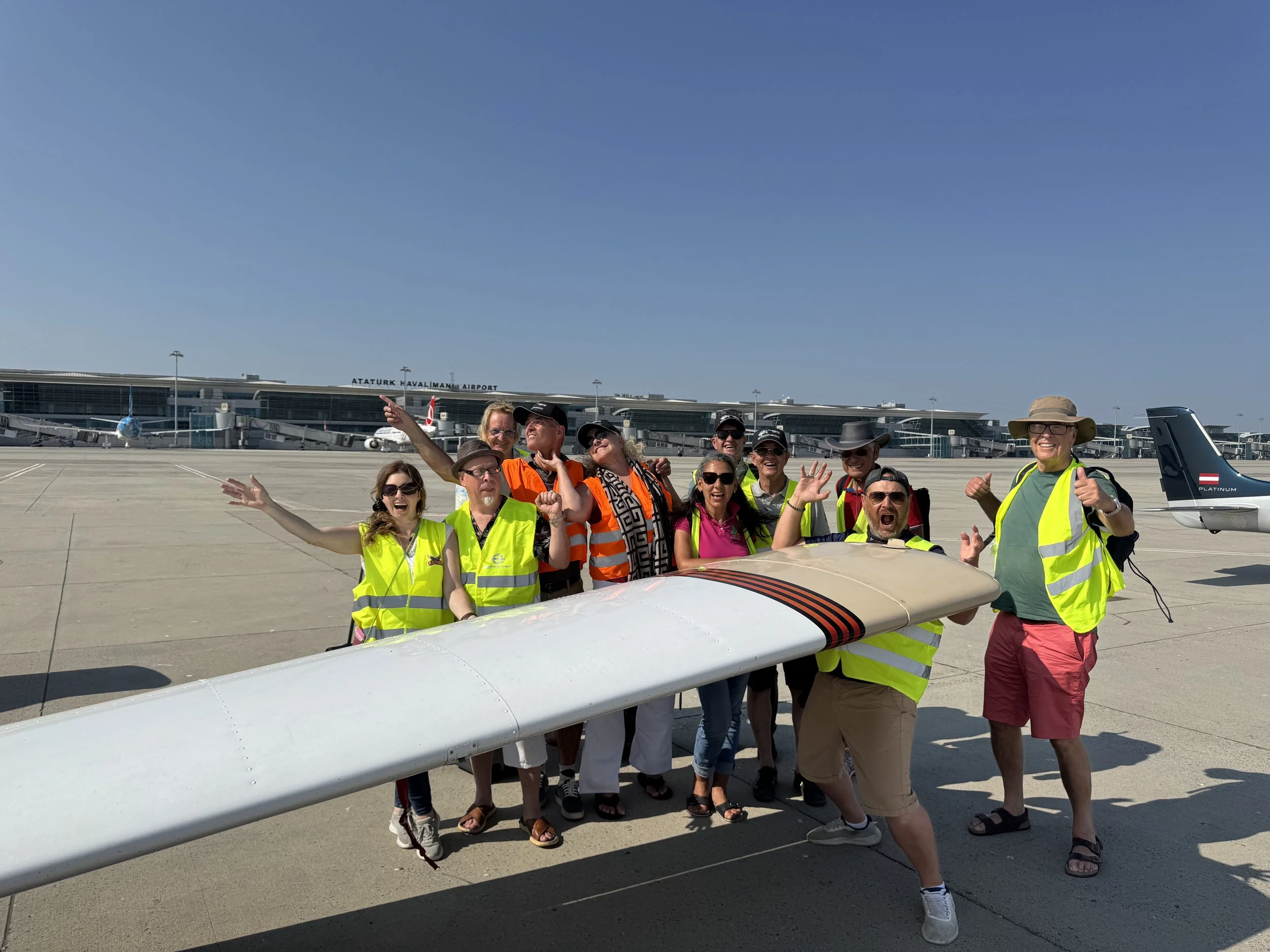 Gruppe von fröhlichen Menschen am Flughafen mit einem Flugzeug im Hintergrund und dem Flughafen-Logo 'Atatürk Flughafen'.
