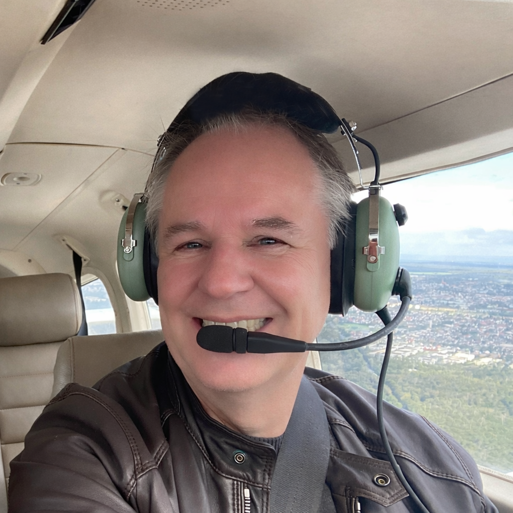 Ein Mann sitzt im Cockpit eines Flugzeugs, trägt Kopfhörer mit Mikrofon und lächelt. Im Hintergrund ist ein Städte- und Landschaftsausblick zu sehen.