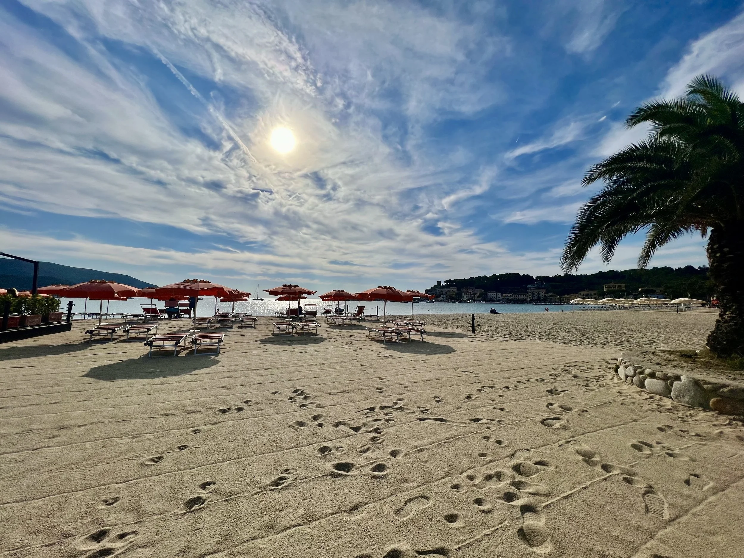 Ein Strand mit Liegestühlen und Sonnenschirmen, Palmen rechts, das Meer im Hintergrund, eine helle Sonne an einem bewölkten Himmel. Sand mit Fußspuren.