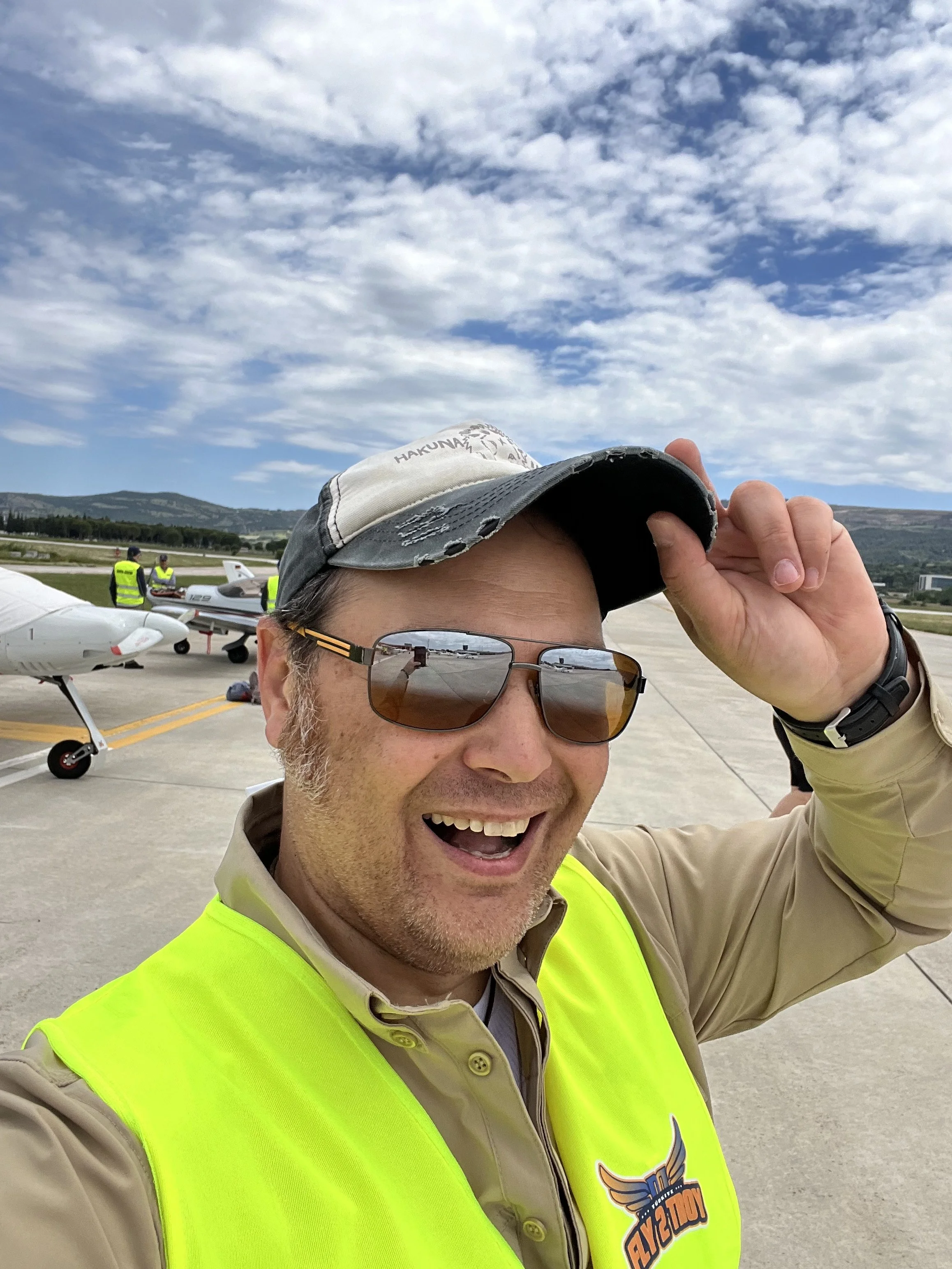 Mann mit Sonnenbrille, Baseballkappe und reflektierenden Sonnenbrille, lächelnd und in einem gelben Sicherheitsweste auf einem Flugplatz, mit einem kleinen Flugzeug im Hintergrund, bei schönem Himmel mit Wolken.