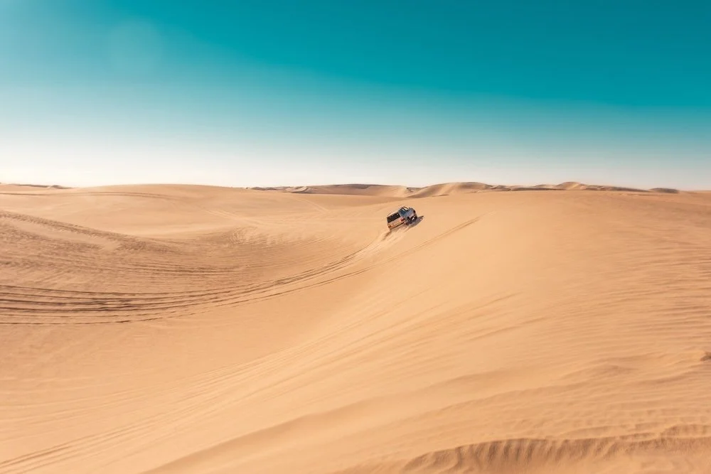 A pickup truck driving on sand dunes in a desert under a clear blue sky.