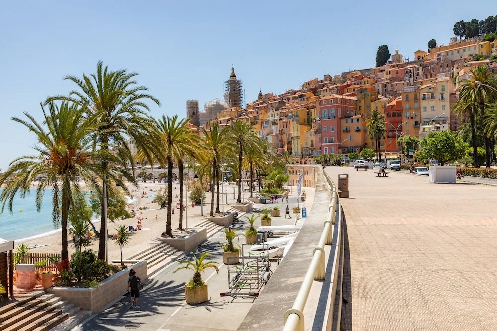 Beachside promenade with palm trees, colorful hillside buildings, and a clear blue sky in a coastal town.