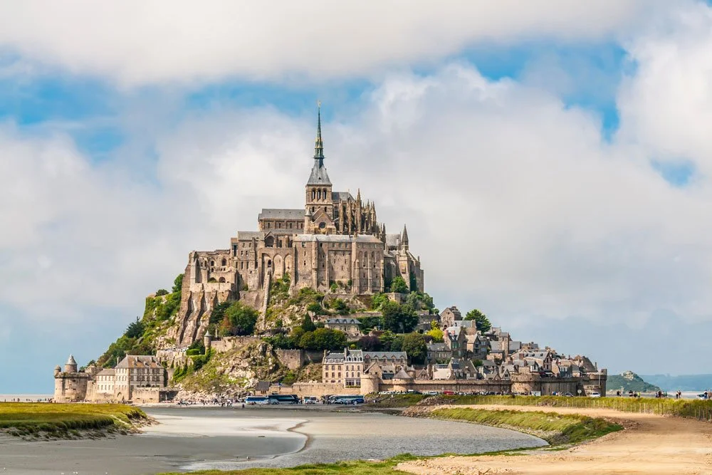 A view of Mont Saint-Michel, a historic island commune in France with a large medieval monastery and surrounding buildings, during daytime with some clouds in the sky.