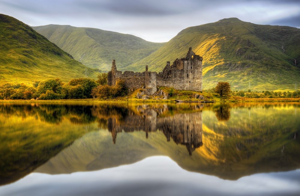 Ancient castle ruins on a lakeshore with mountain range behind, reflection in the water.