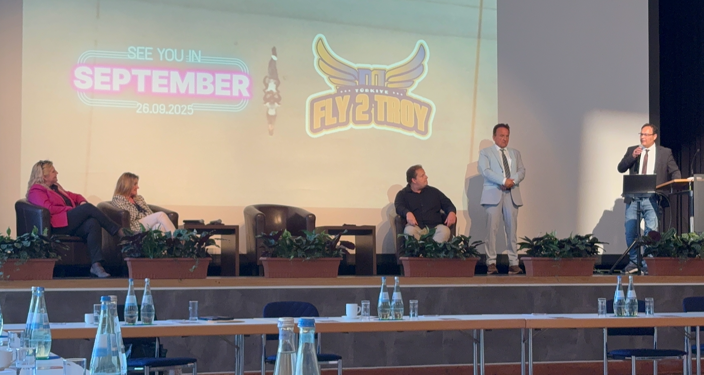 Vier Personen sitzen auf einer Bühne, während eine Person rechts spricht. Im Hintergrund ist ein Bildschirm mit dem Logo "Fly2Troy" und dem Text "See you in September 26.09.2025" zu sehen. Im Vordergrund stehen Tische mit Wasserflaschen.