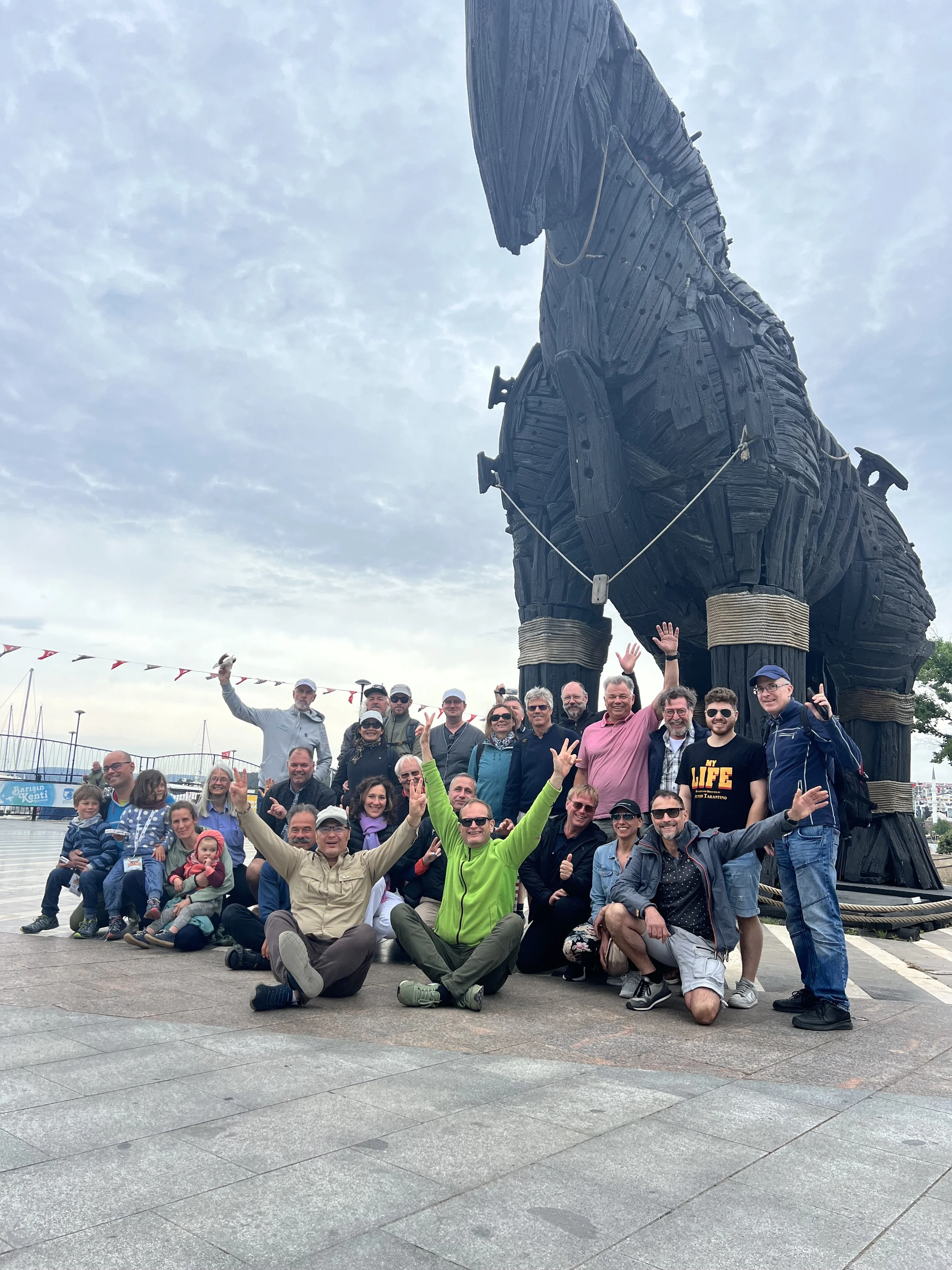 Gruppenfoto von Touristen vor einer großen Statue von einem Pferd aus Holz. Die Gruppe besteht aus Erwachsenen und Kindern, die lächelnd in die Kamera schauen und einige winken. Das Wetter ist bewölkt.