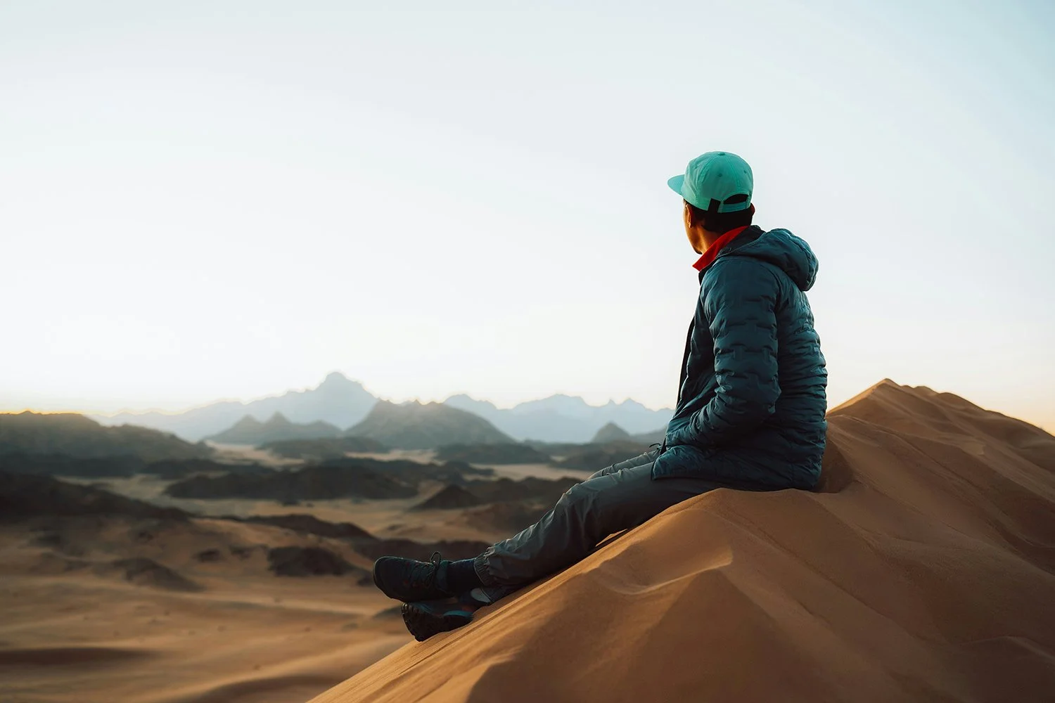 man in a teal hat sits upon a sand dune and looks out to the mountains in the distance