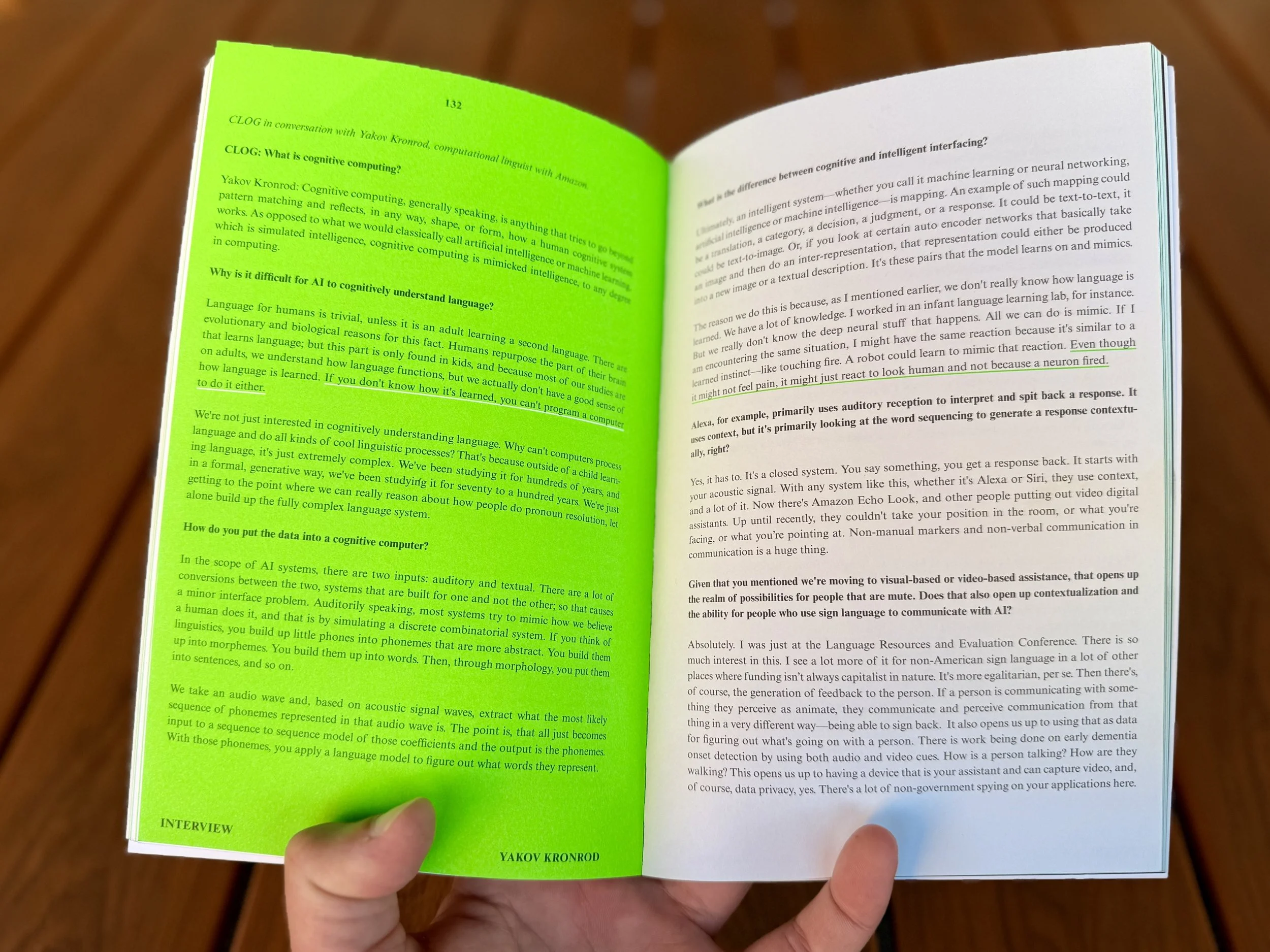 Open book with highlighted green text on left page and black text on right page, held in someone's left hand over a wooden floor.