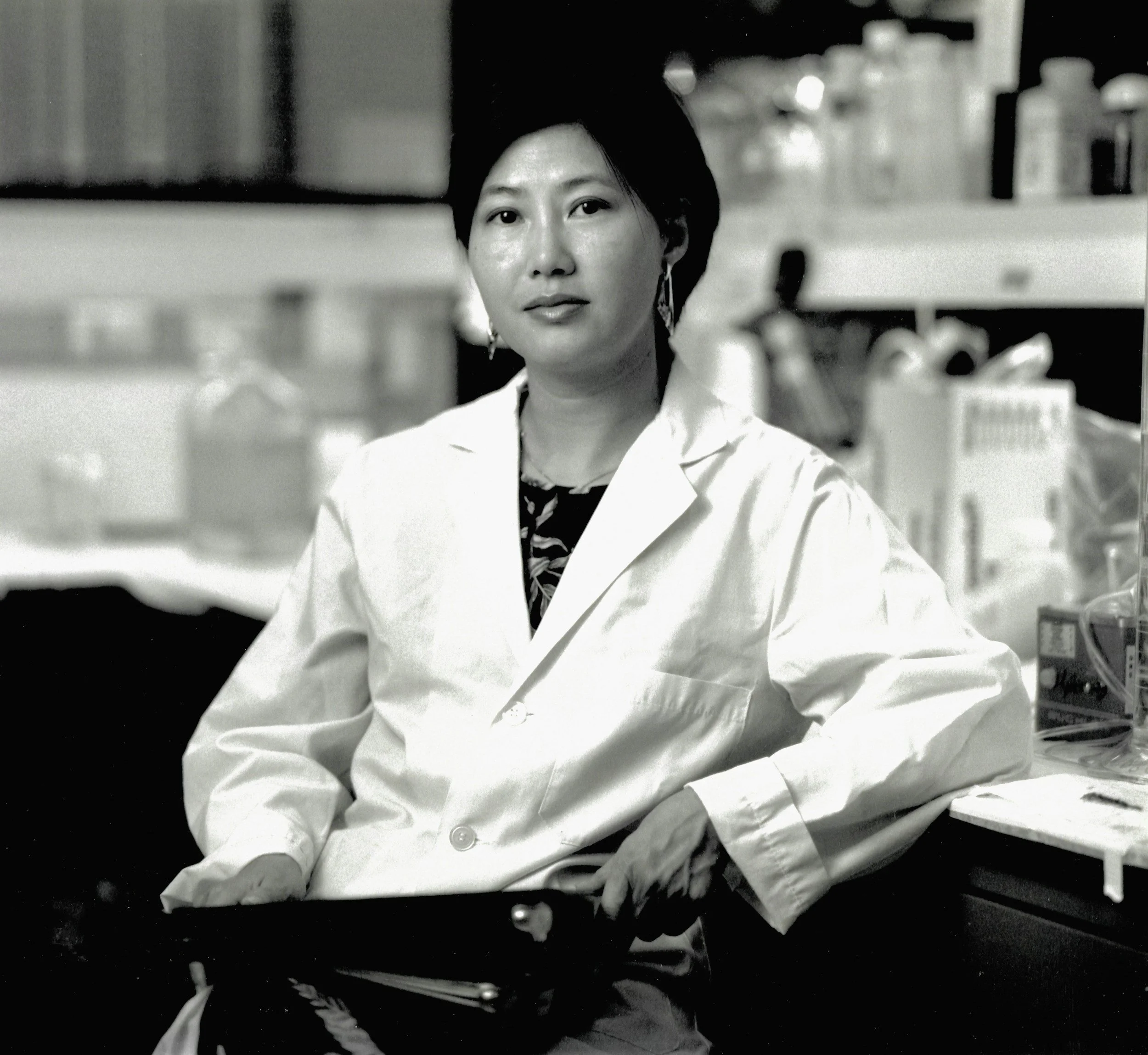 A woman in a lab coat standing in a laboratory environment with shelves of bottles and lab equipment behind her.