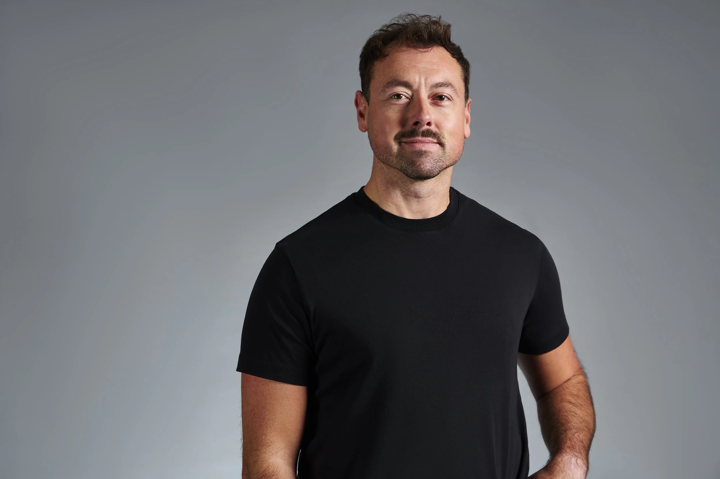 A man with short brown hair, slight beard, wearing a black t-shirt, stands against a plain gray background.