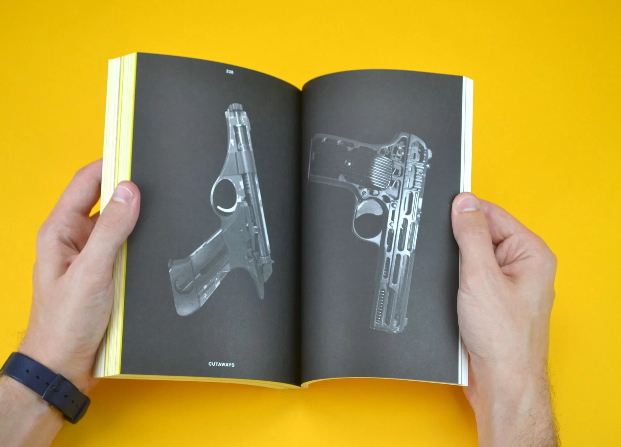 Open magazine showing black and white images of firearm components on dark background, held by person with light skin and black watch band against yellow background.