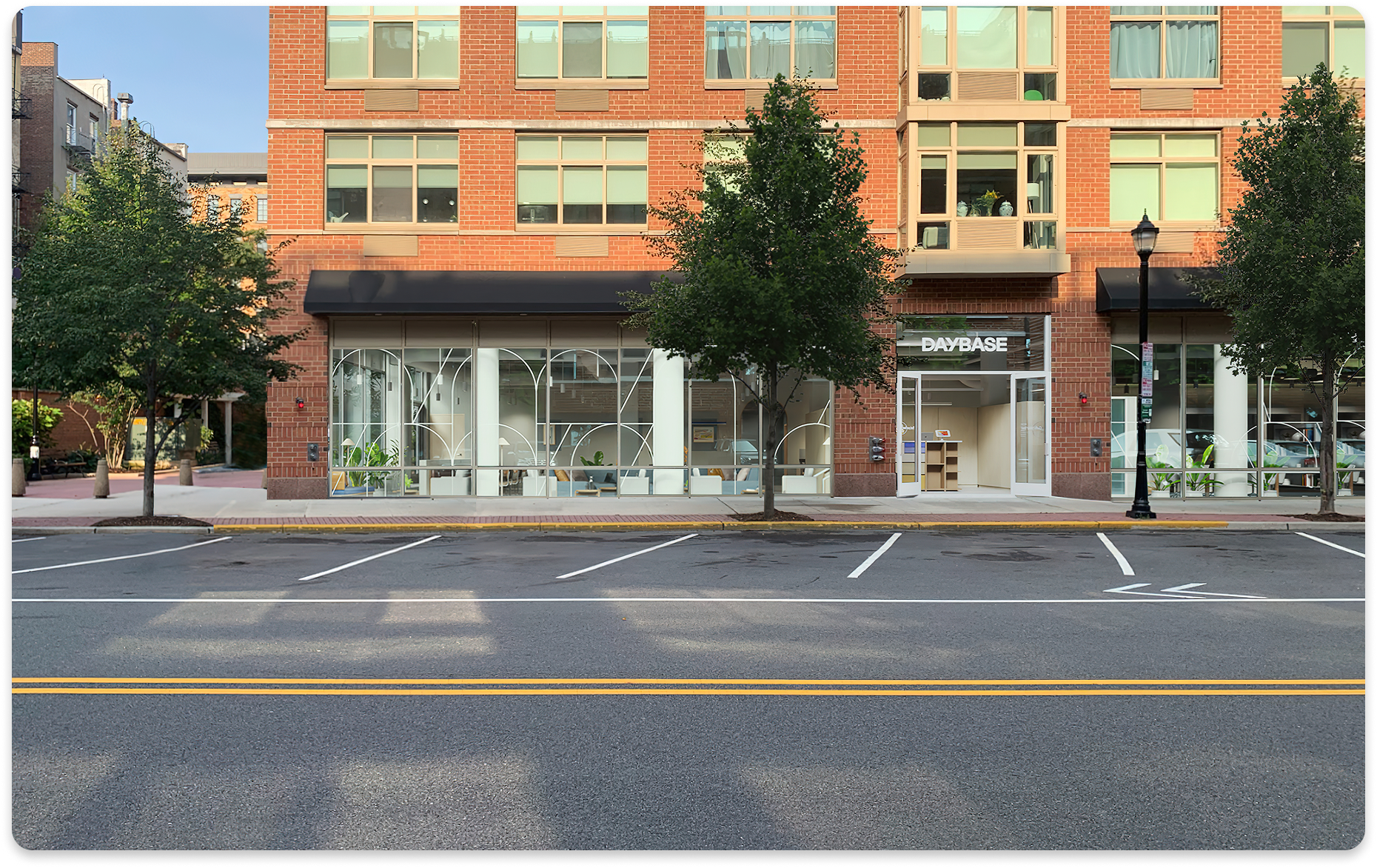 Empty street with parking lot, trees, and a brick building with storefronts, including one named DAYBASE, large windows, and black awnings.