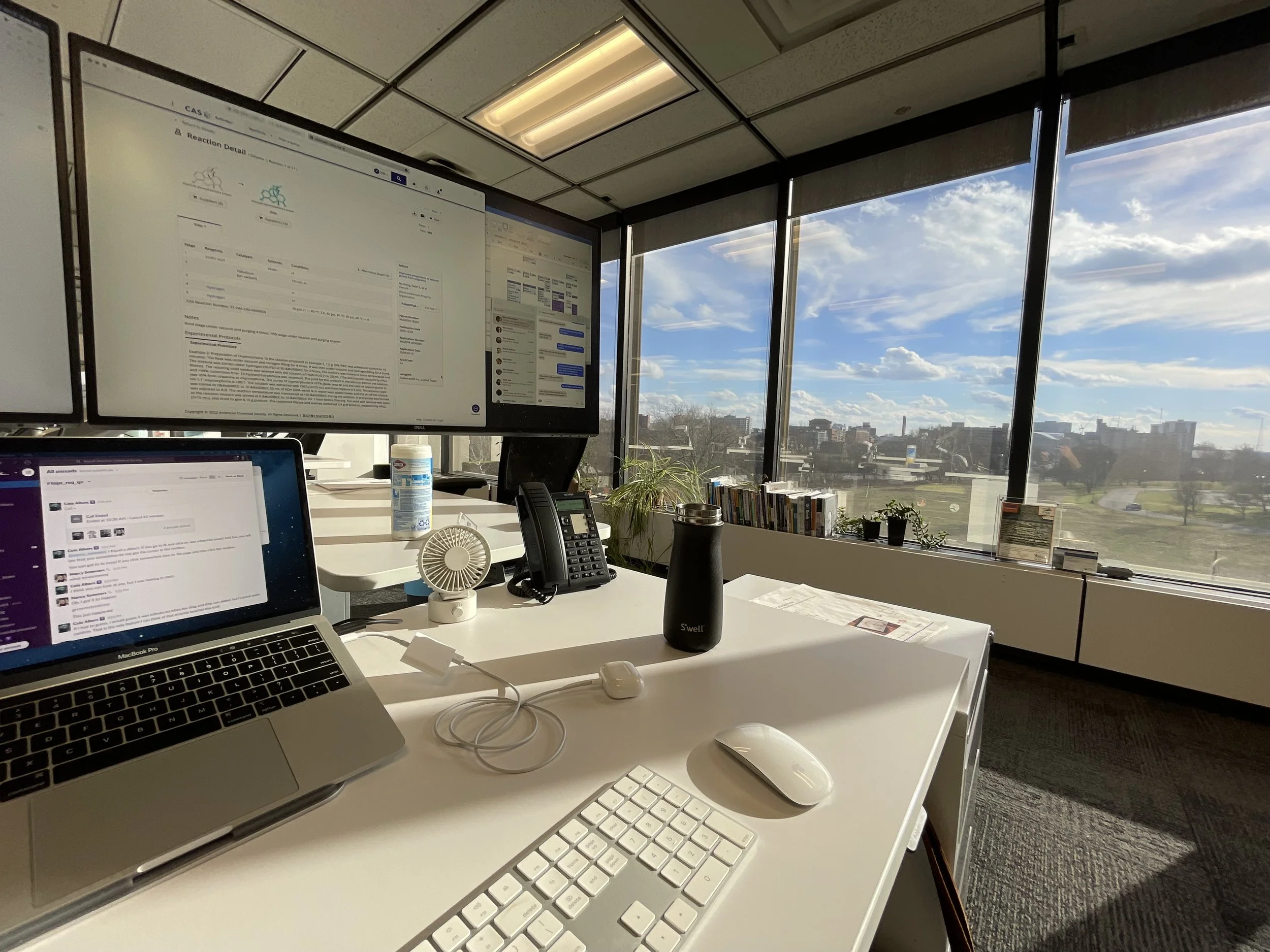 Office workspace with a white desk, a laptop, dual monitors, a keyboard, a mouse, a black water bottle, a telephone, a small fan, and plants by large windows showing a cityscape and blue sky.