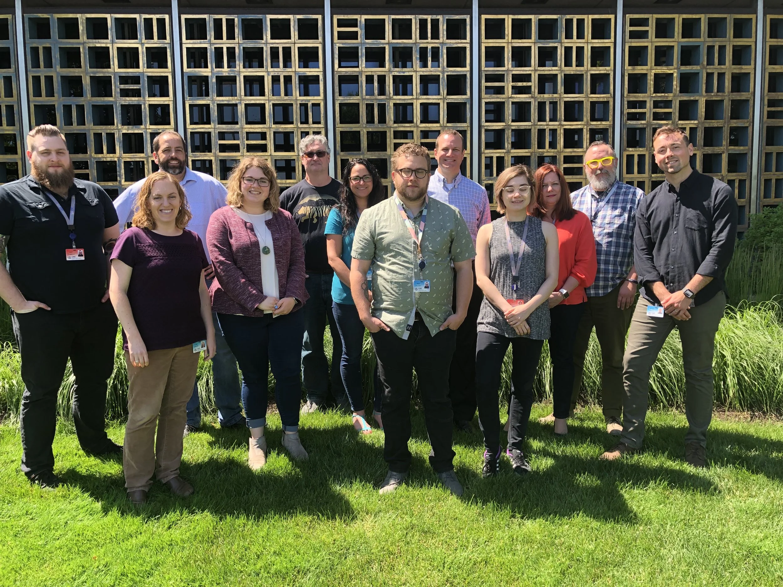 Group of fifteen diverse people standing outdoors on a sunny day in front of a modern wooden building with green grass and plants.