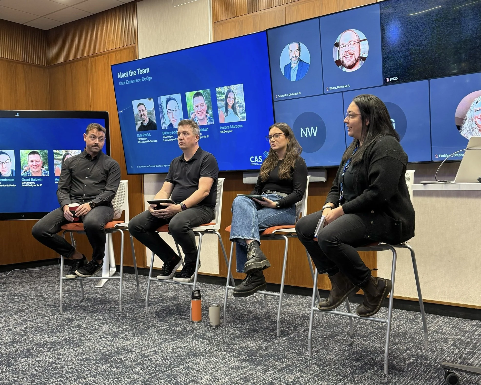 A panel discussion with four individuals seated on high chairs in front of a large digital screen displaying a team introduction for user experience design. The room has wood-paneled walls and a carpeted floor. The panelists are holding tablets or notebooks, and there are water bottles on the floor.