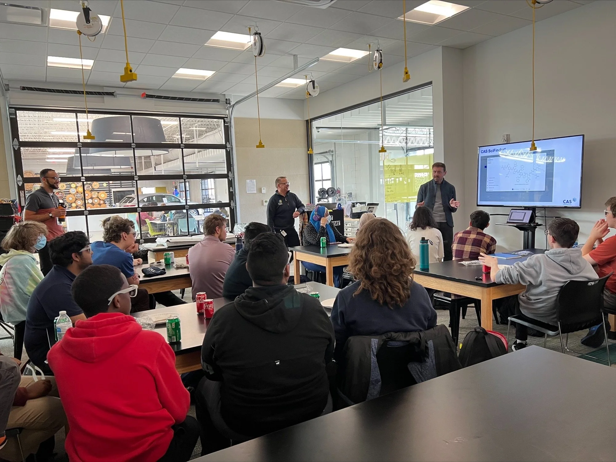 A group of students attending a classroom presentation, with a male instructor presenting on a large screen, inside a modern educational space with a glass garage door and ceiling-mounted projectors.