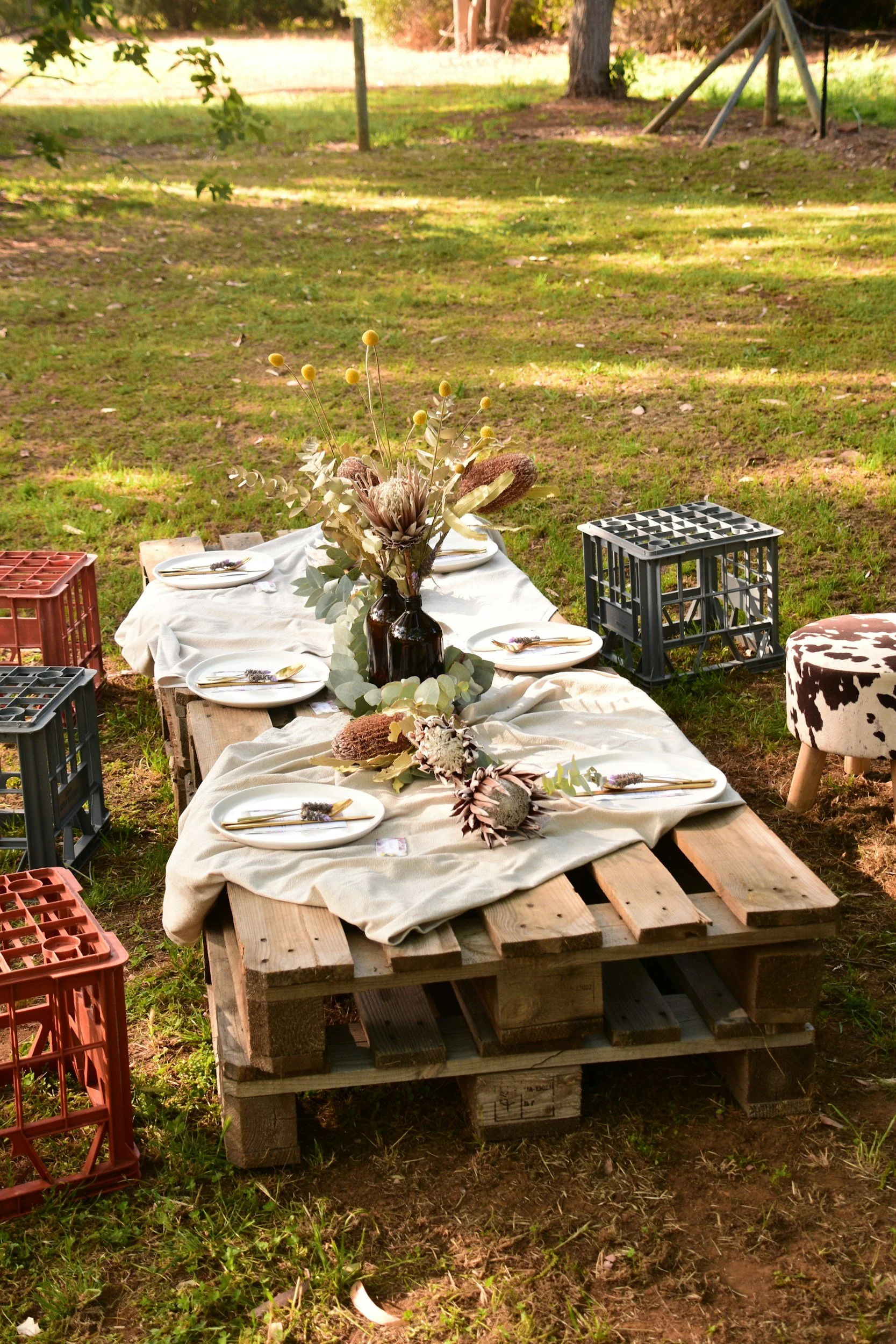 Outdoor dining table made from wooden pallets with a white tablecloth, decorated with a bouquet of dried flowers in a dark vase, set with white plates, gold cutlery, and decorative items, surrounded by milk crates and a stool, in a grassy yard with trees and a swing in the background.