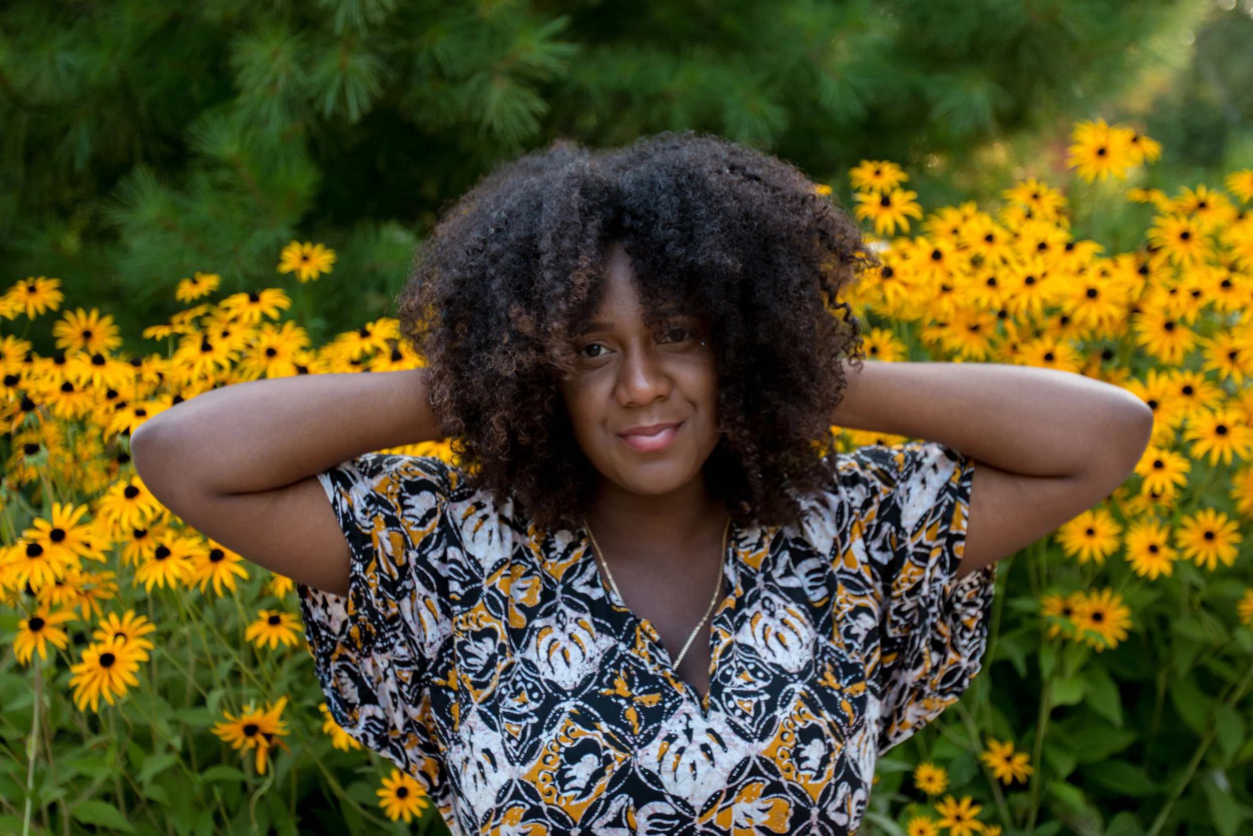A black woman with dark curly hair smiling with her hands behind her head, standing in front of yellow flowers and green foliage.