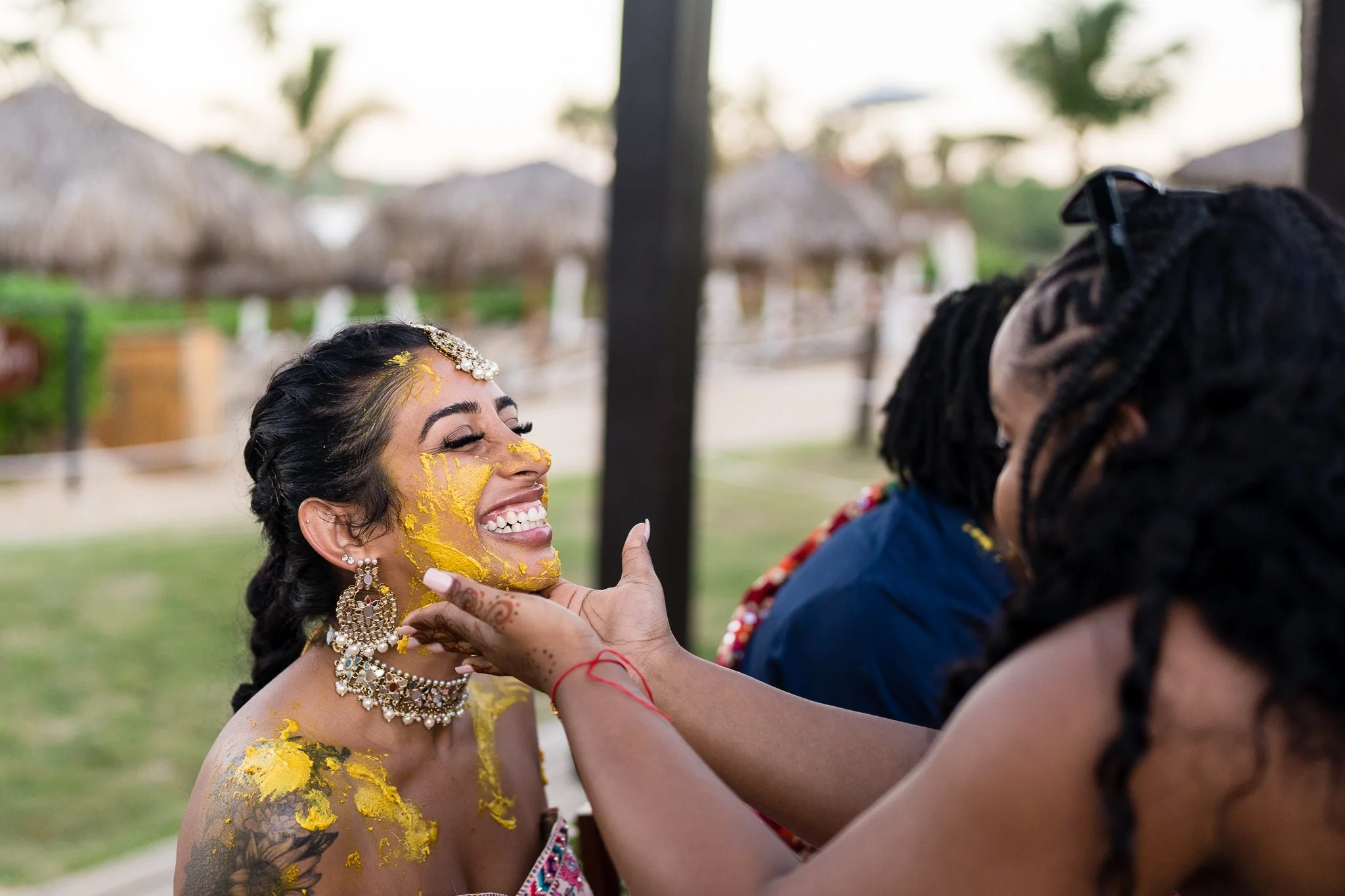 A woman with traditional Indian jewelry has yellow powder applied to her face and shoulders during a Maiyan celebration