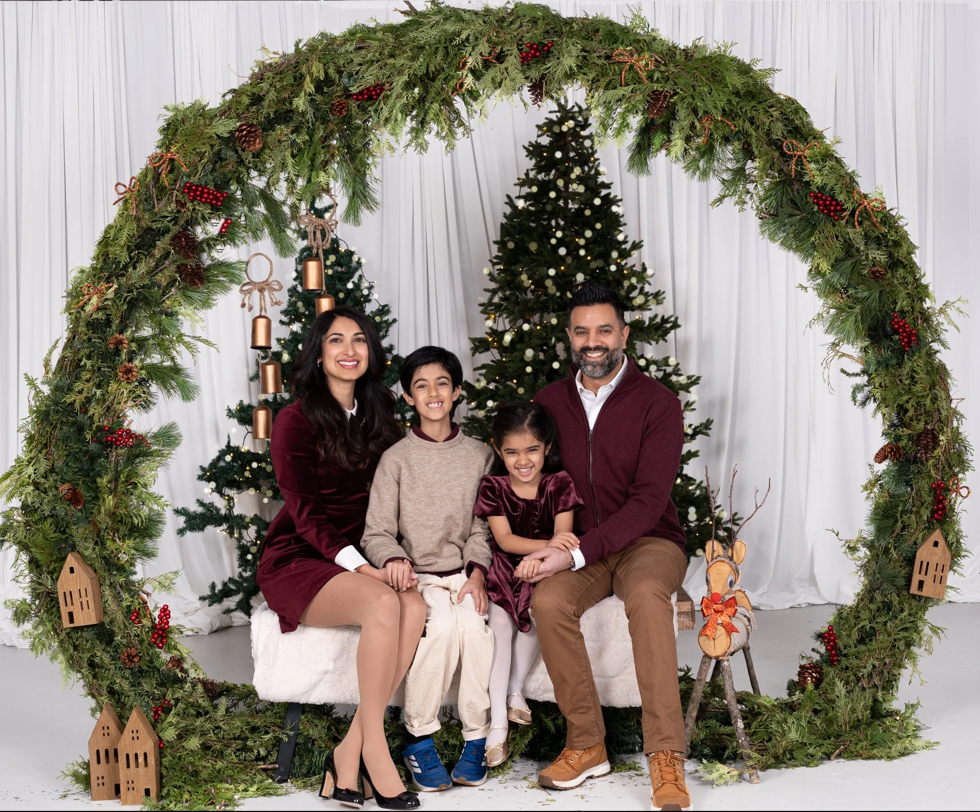 Family of four people posing under a Christmas arch decorated with greenery, pinecones, berries, and ribbons, with Christmas trees in the background.