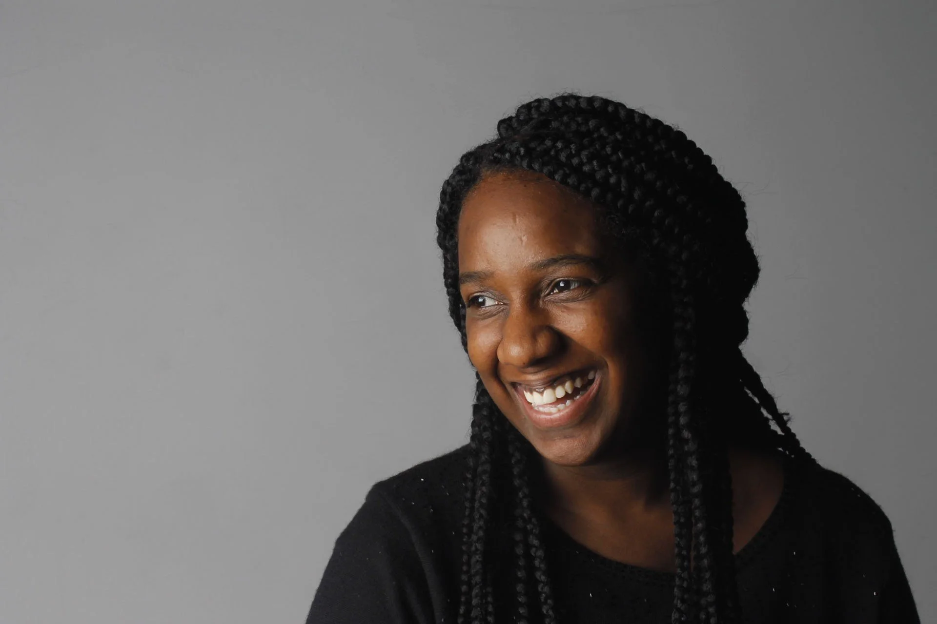 A smiling woman with dark skin and braided hair, wearing a black shirt, in front of a gray background.