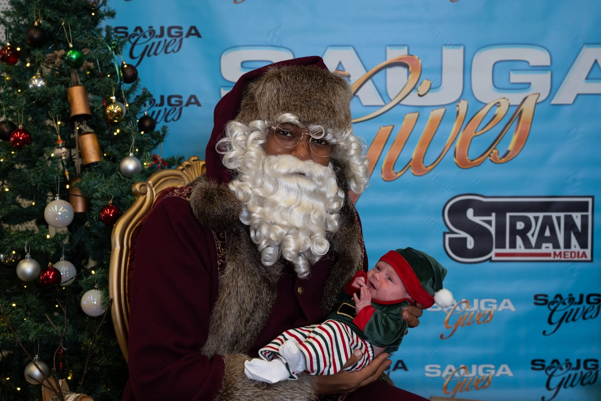 Santa sitting in front of a Christmas tree holding a small infant at the 2025 Sauga Gives Christmas event