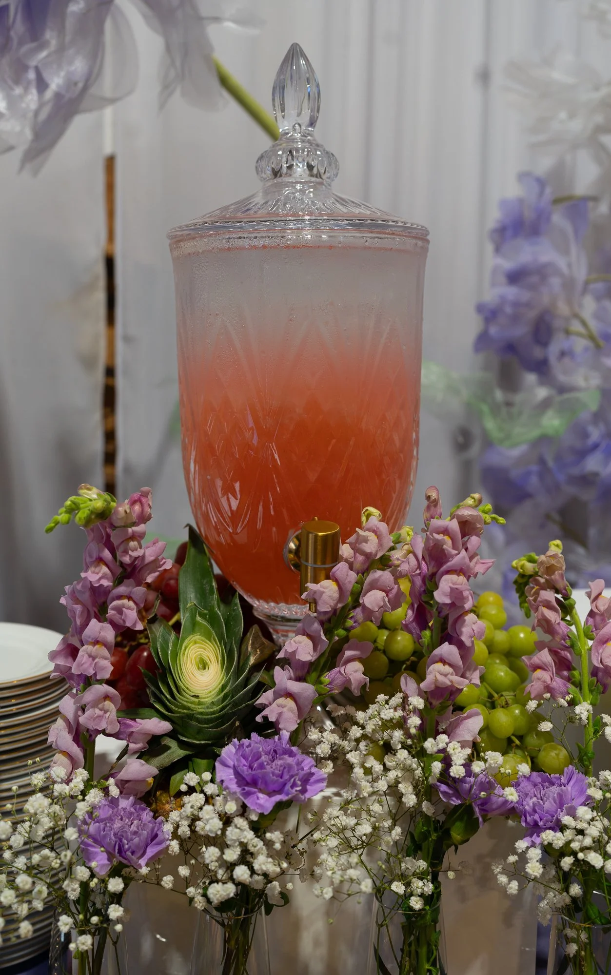 Birthday party decorations. A large glass dispenser  filled with a pinkish liquid, surrounded by colourful flowers around it.