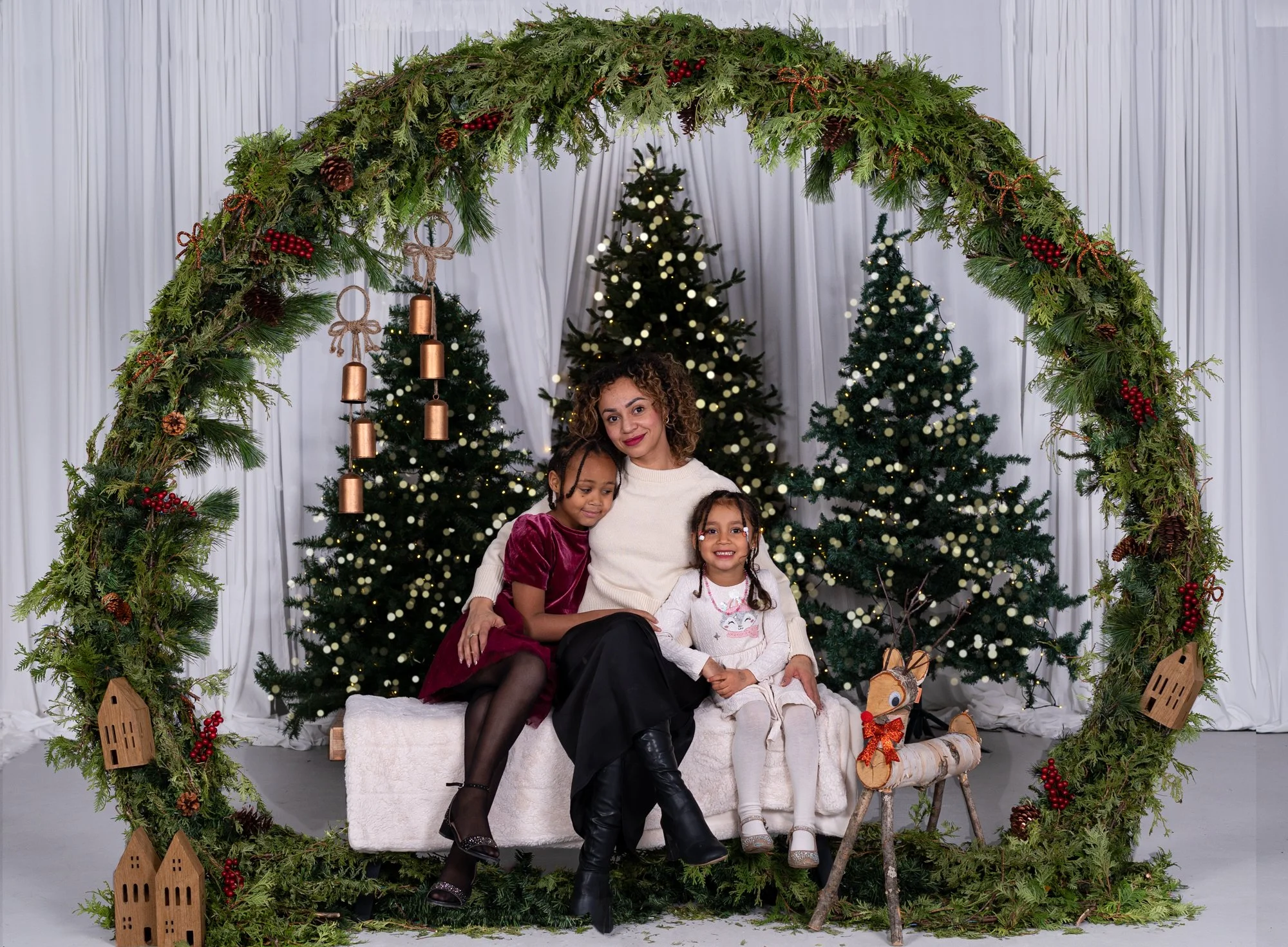 A mother and her two young girls sitting within a large circular green garland arch surrounded by decorated holiday decorations and Christmas trees.