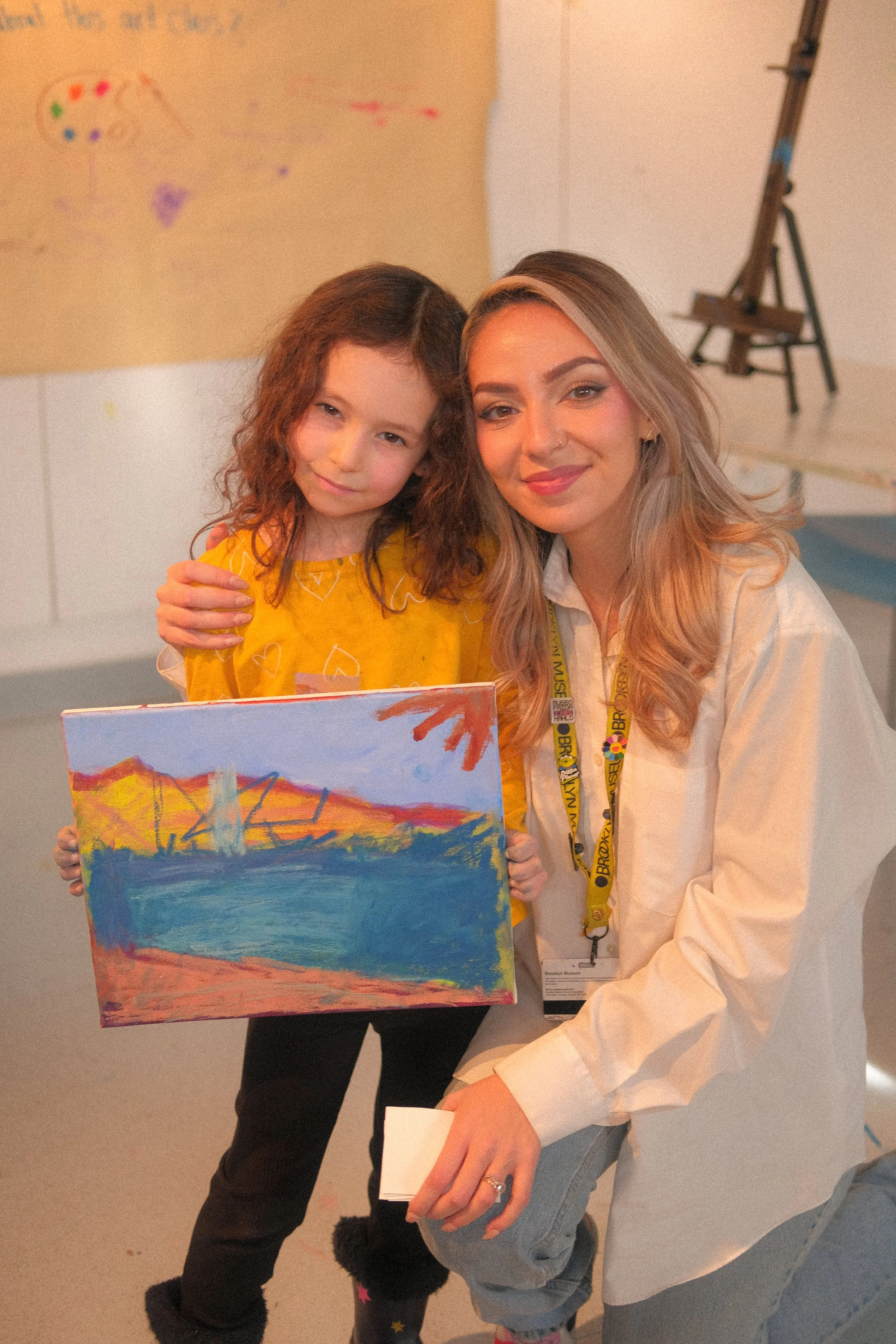 A young girl in a yellow shirt holding a colorful painting, standing next to an adult woman with long blonde hair, both smiling, in an indoor setting with a beige wall and an easel in the background.