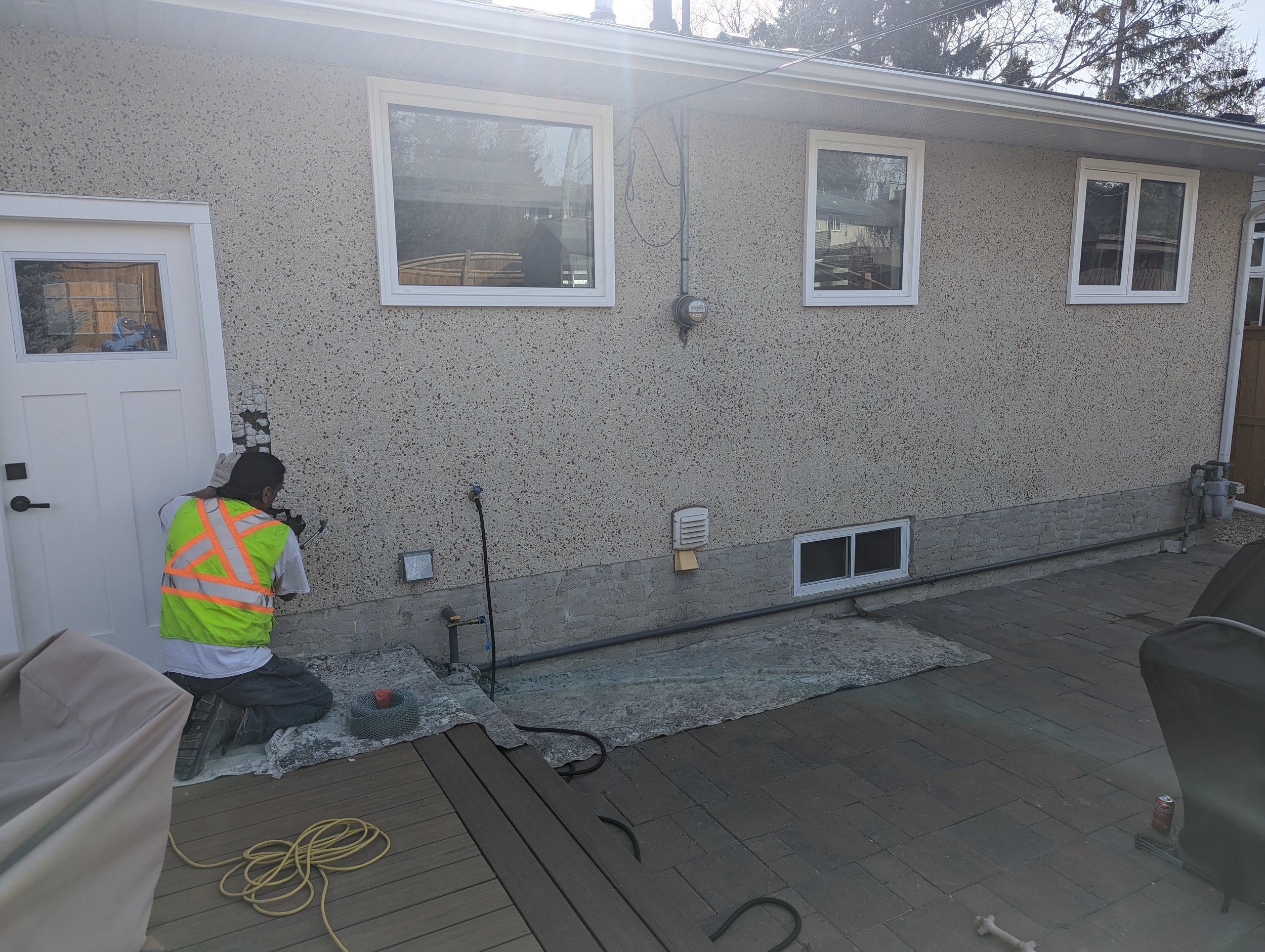A person kneeling on a protective sheet next to a house wall, working on an outdoor electrical or plumbing fixture. The house has a stucco exterior with three windows and a white door. The area is paved with bricks, and various tools and materials are visible.