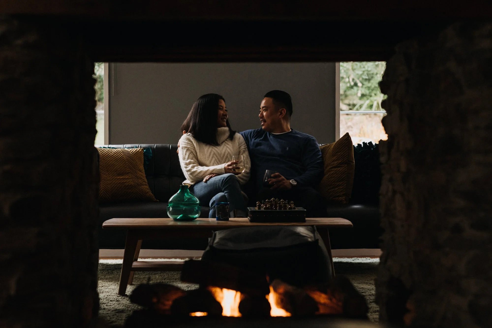 A couple sitting on a black sofa, smiling and looking at each other, holding drinks, behind a small table with green vase and chess set, seen through a fireplace opening.