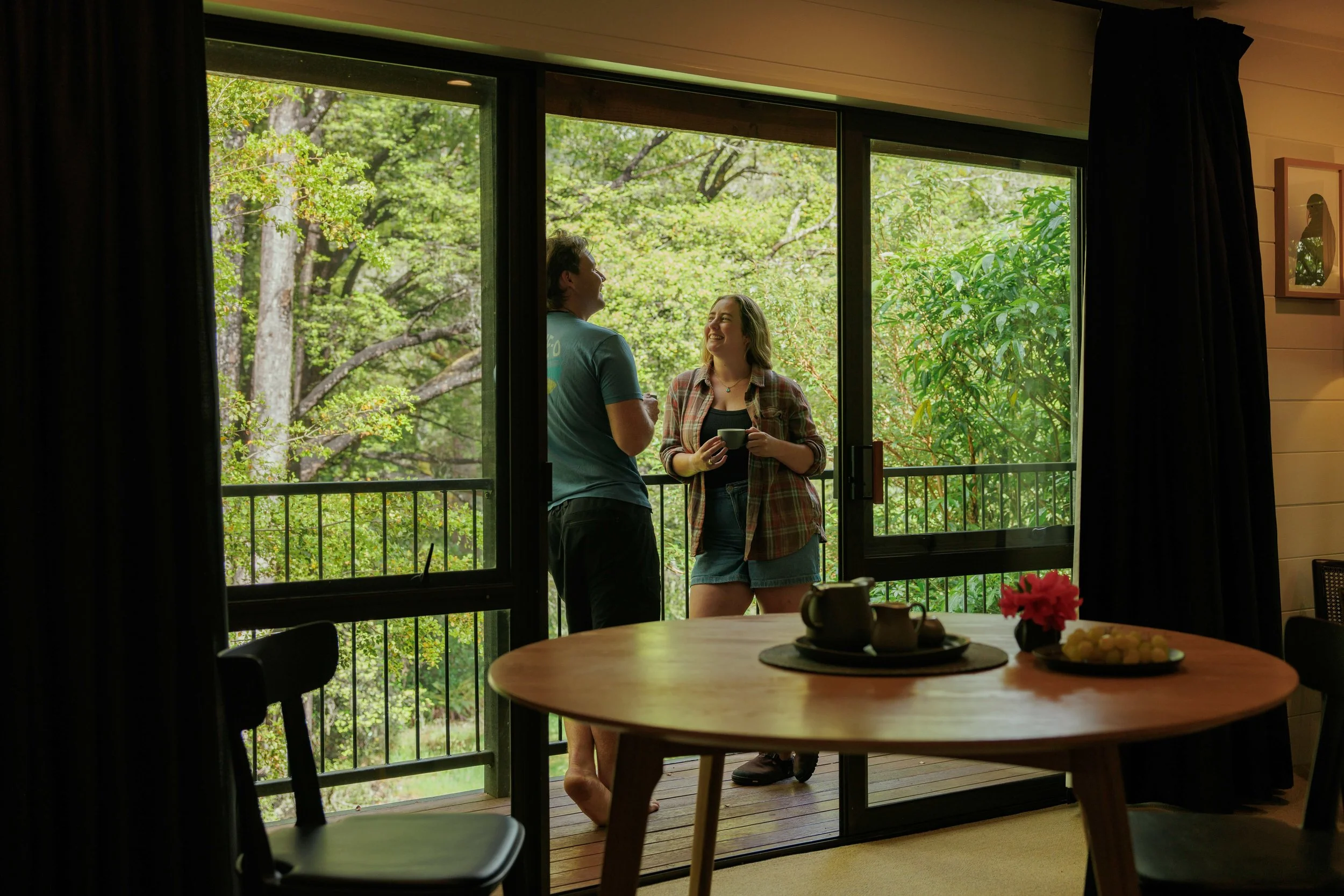 A person stands barefoot on a balcony, looking out over lush green trees and forested landscape.